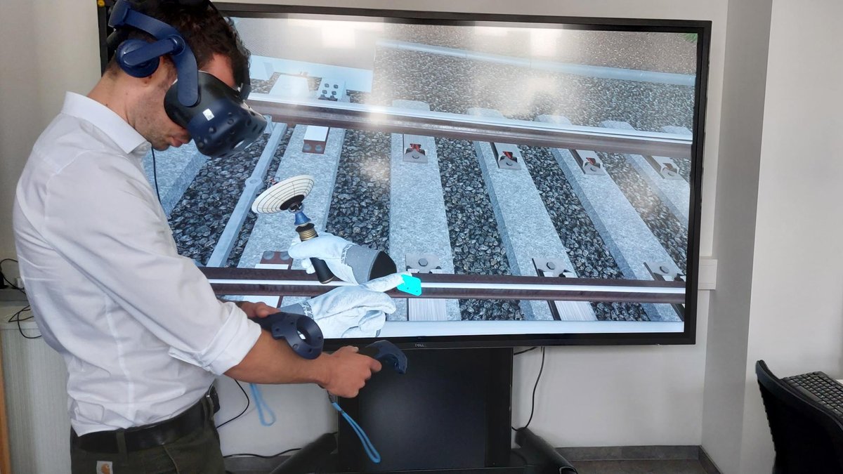 Ein Mann trägt eine VR-Brille, auf einem Bildschirm im Hintergrund sind Bahngleise abgebildet.