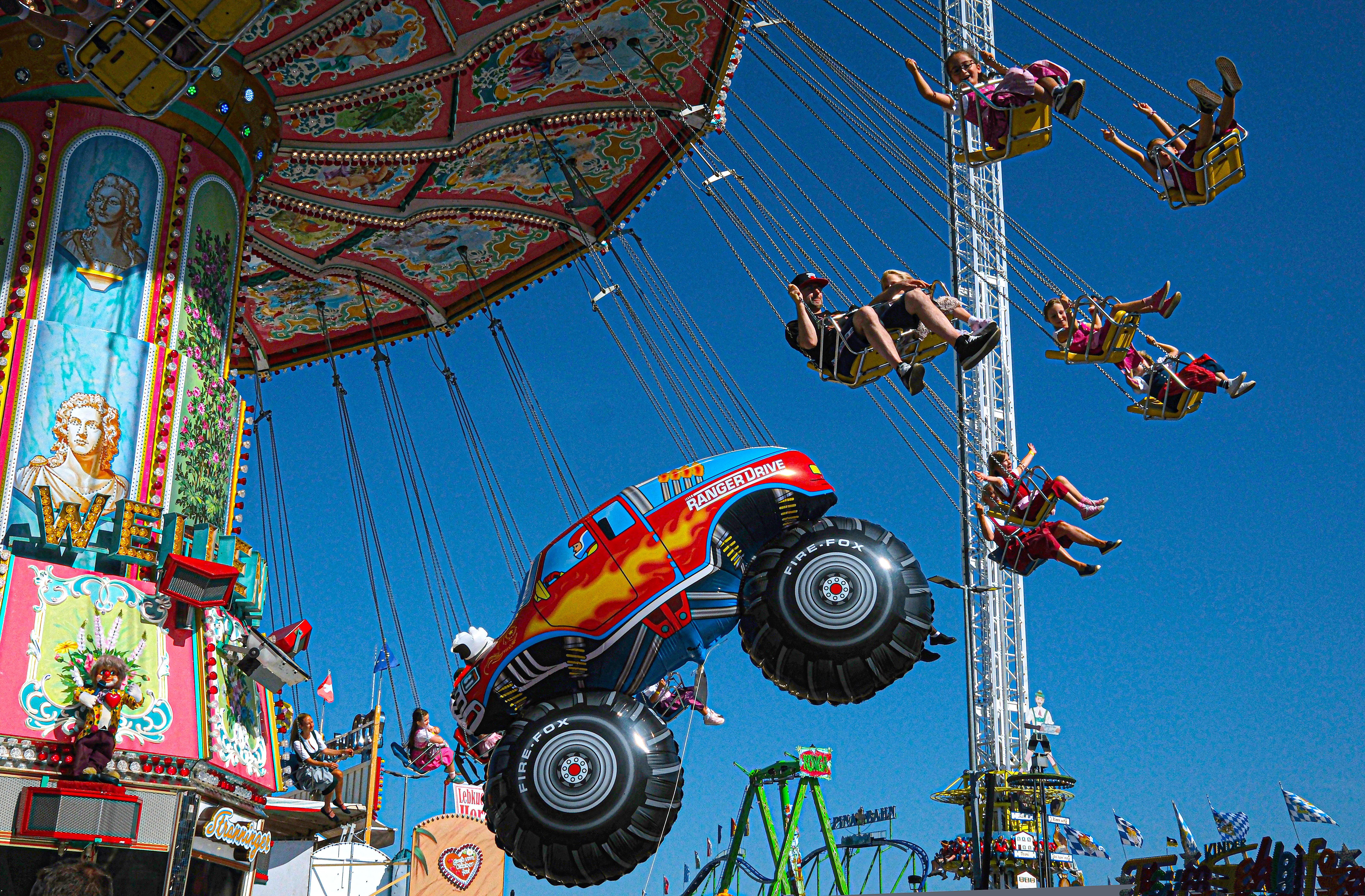 Menschen im Kettenkarussell, dahinter ein "Freefall Tower" und ein Luftballon in Form eines Trucks.