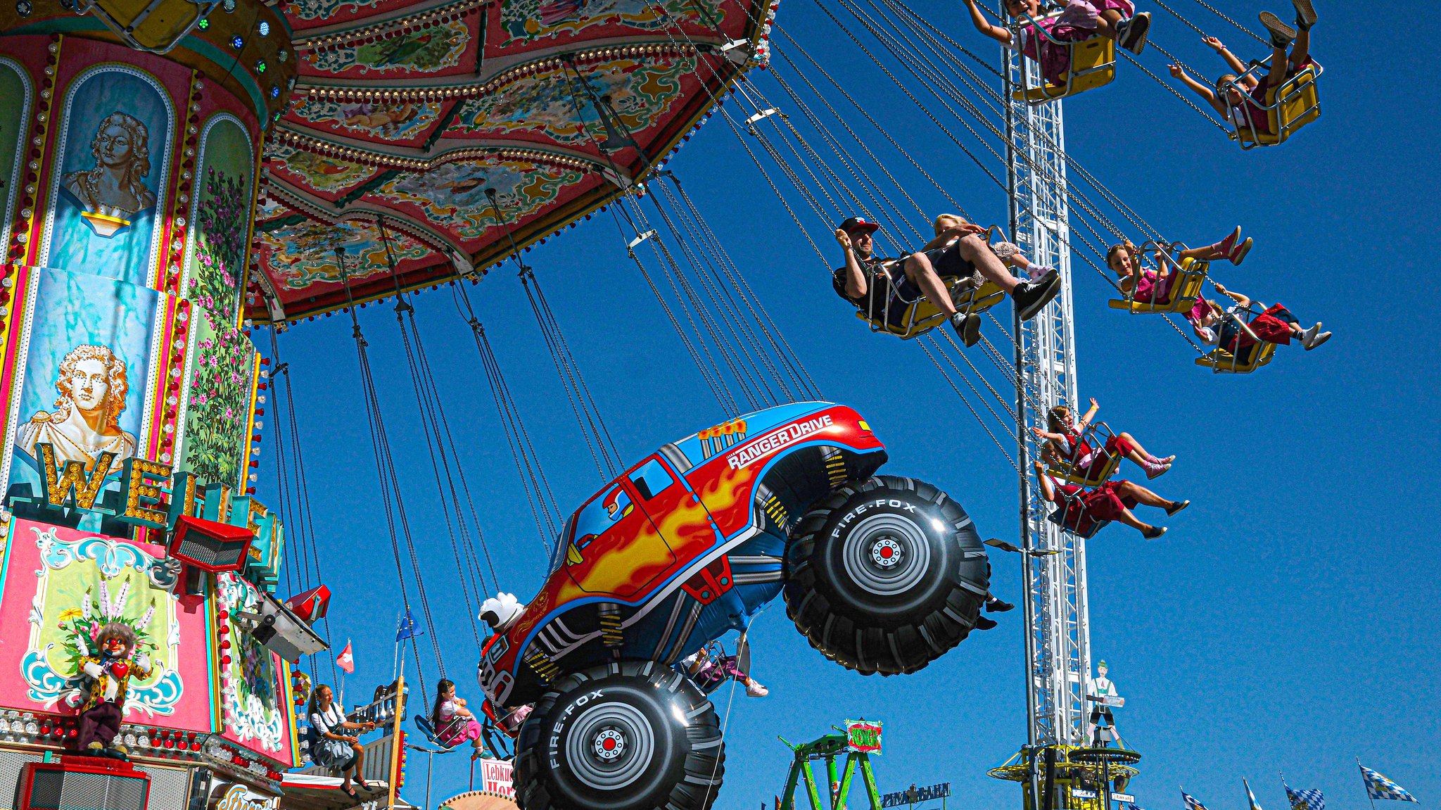Menschen im Kettenkarussell, dahinter ein "Freefall Tower" und ein Luftballon in Form eines Trucks.