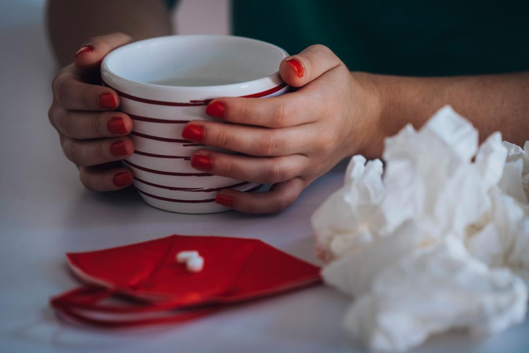 Hände umschließen eine Tasse. Daneben liegt eine halbierte Tablette auf einer roten FFP2-Maske und gebrauchte Taschentücher.