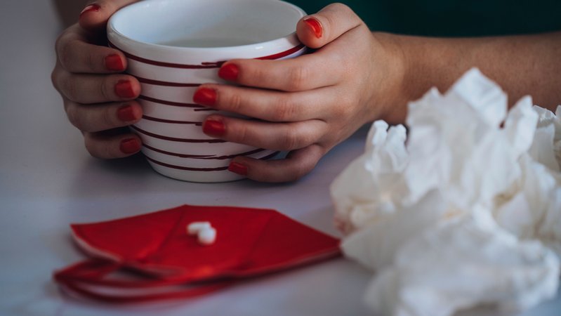Hände umschließen eine Tasse. Daneben liegt eine halbierte Tablette auf einer roten FFP2-Maske und gebrauchte Taschentücher. | Bild: BR/Raphael Kast Hände umschließen eine Tasse. Daneben liegt eine halbierte Tablette auf einer roten FFP2-Maske und gebrauchte Taschentücher.