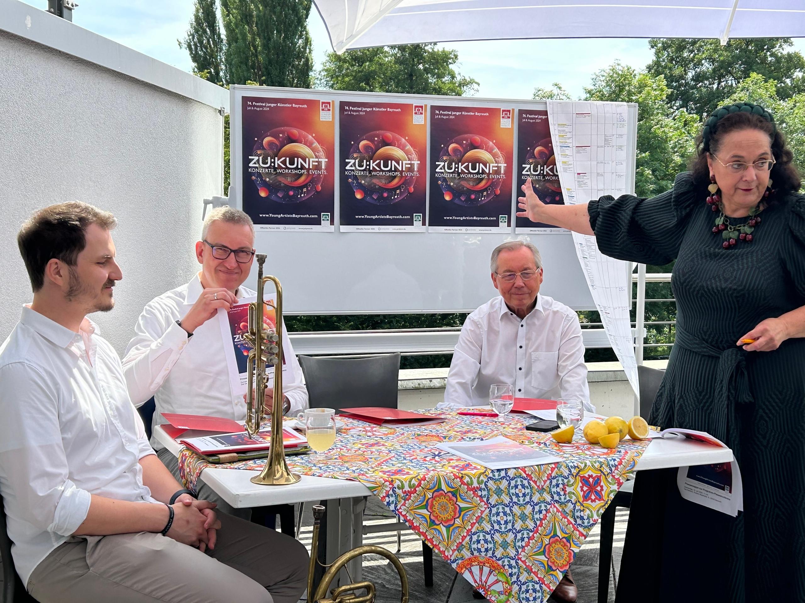 Musiker sitzen an einem Tisch, auf dem eine Trompete steht, dahinter ein Wand mit drei Plakaten, auf denen "Zukunft" zu lesen ist.