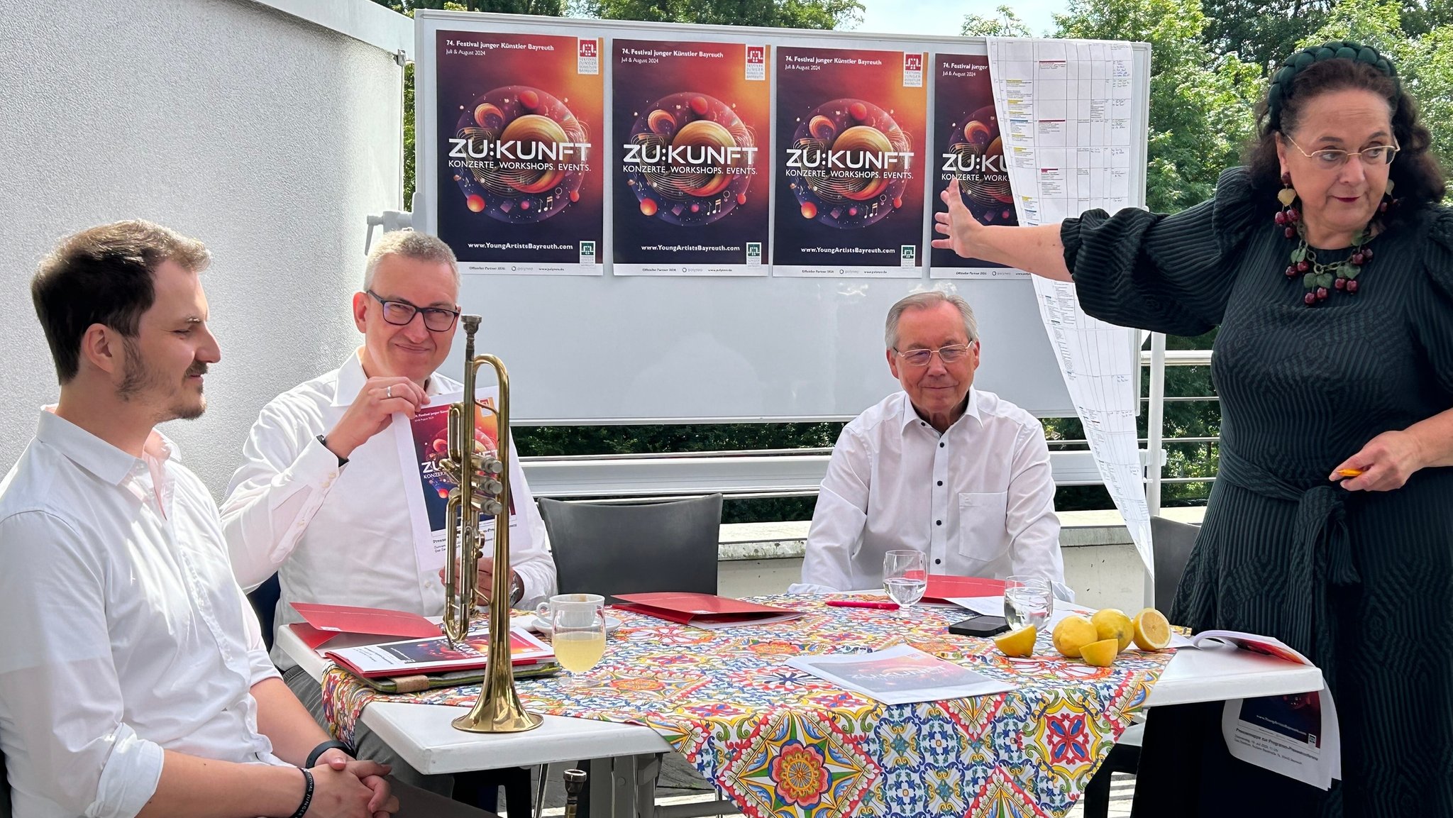 Musiker sitzen an einem Tisch, auf dem eine Trompete steht, dahinter ein Wand mit drei Plakaten, auf denen "Zukunft" zu lesen ist.