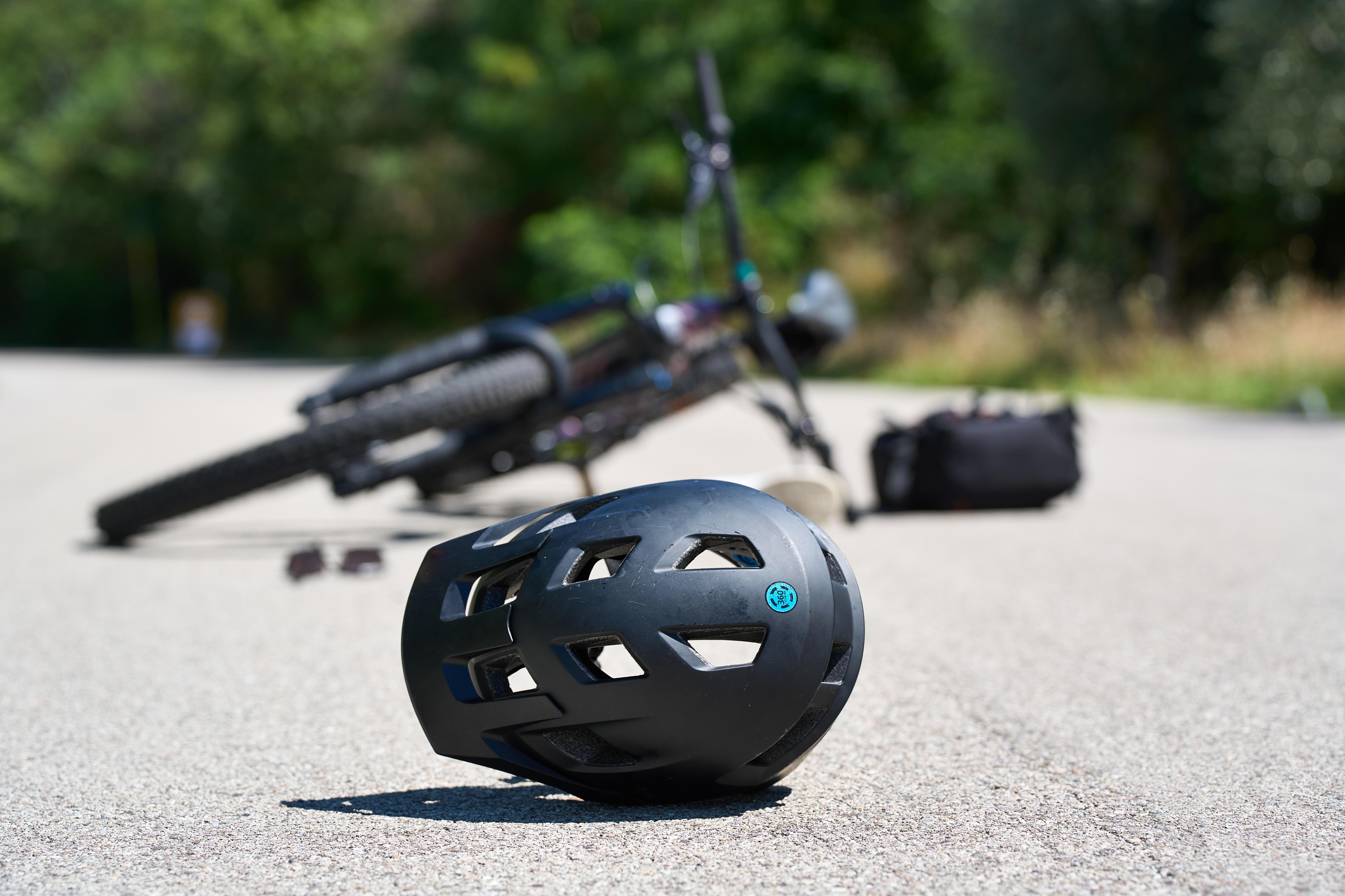 Symbolbild für einen Fahrradunfall: Ein Helm liegt verlassen auf der Straße, im Hintergrund ein gestürztes Fahrrad 