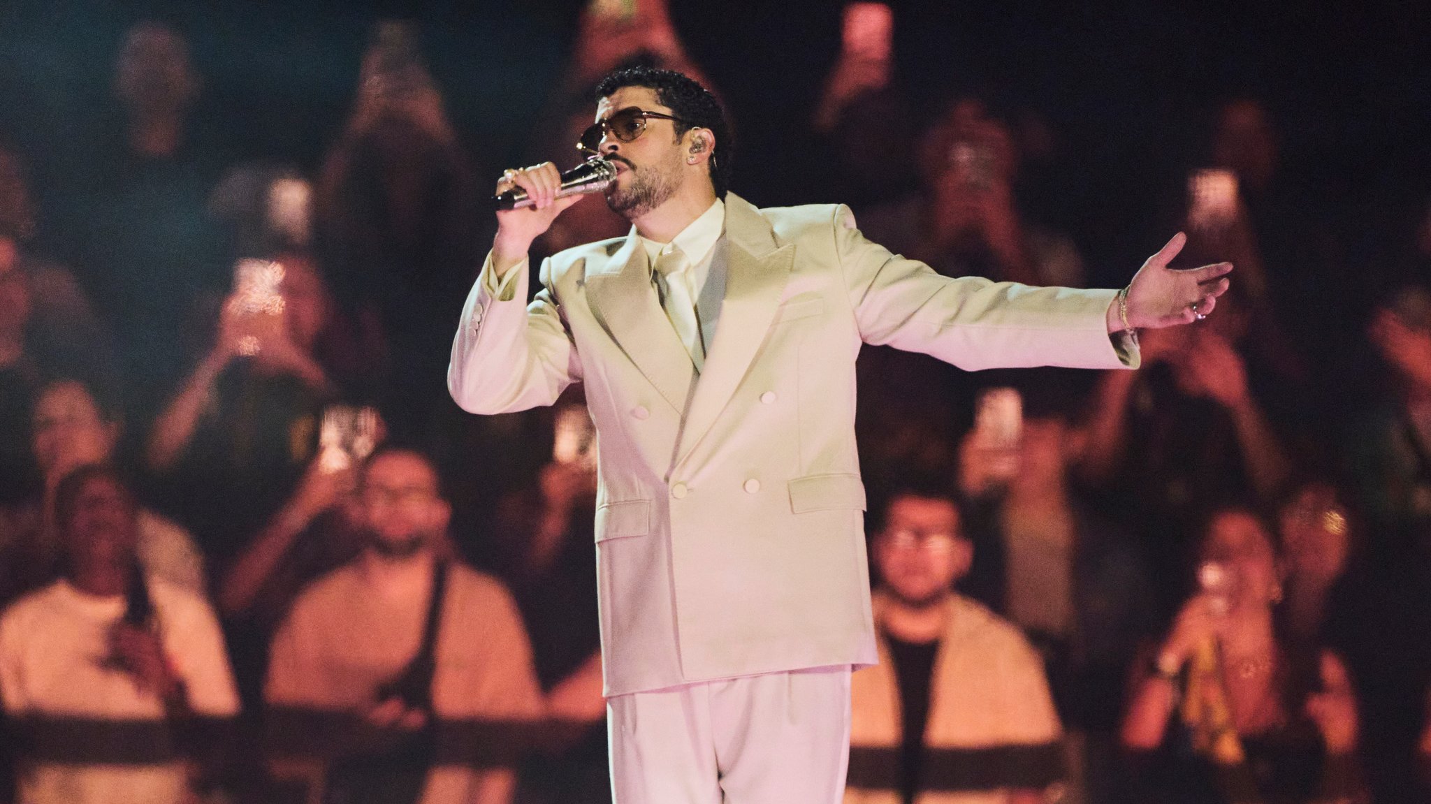 Auf dem Bild ist der Künstler Bad Bunny in einem weißen Anzug, bei seinem Auftritt in Buenos Aires (Argentinien) Anfang Februar zu sehen. Er hält ein Mikrofon in seiner Hand und singt. 