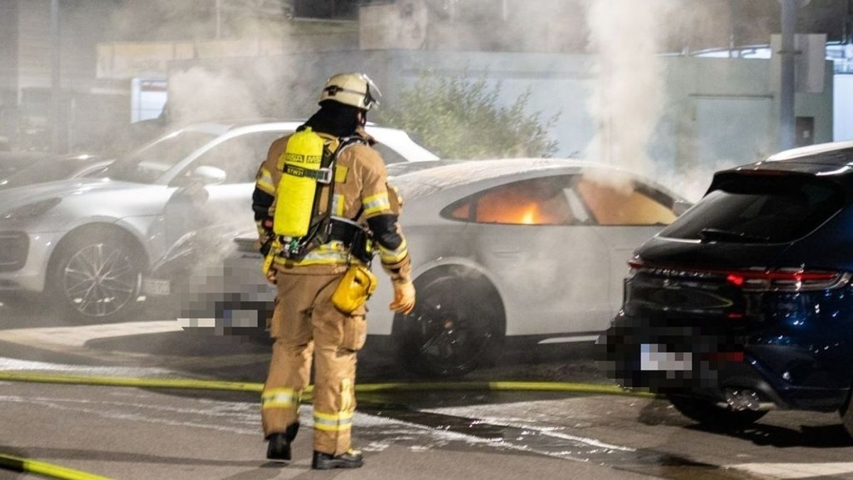 Ein E-Auto brennt. Ein Feuerwehrmann steht davor. 