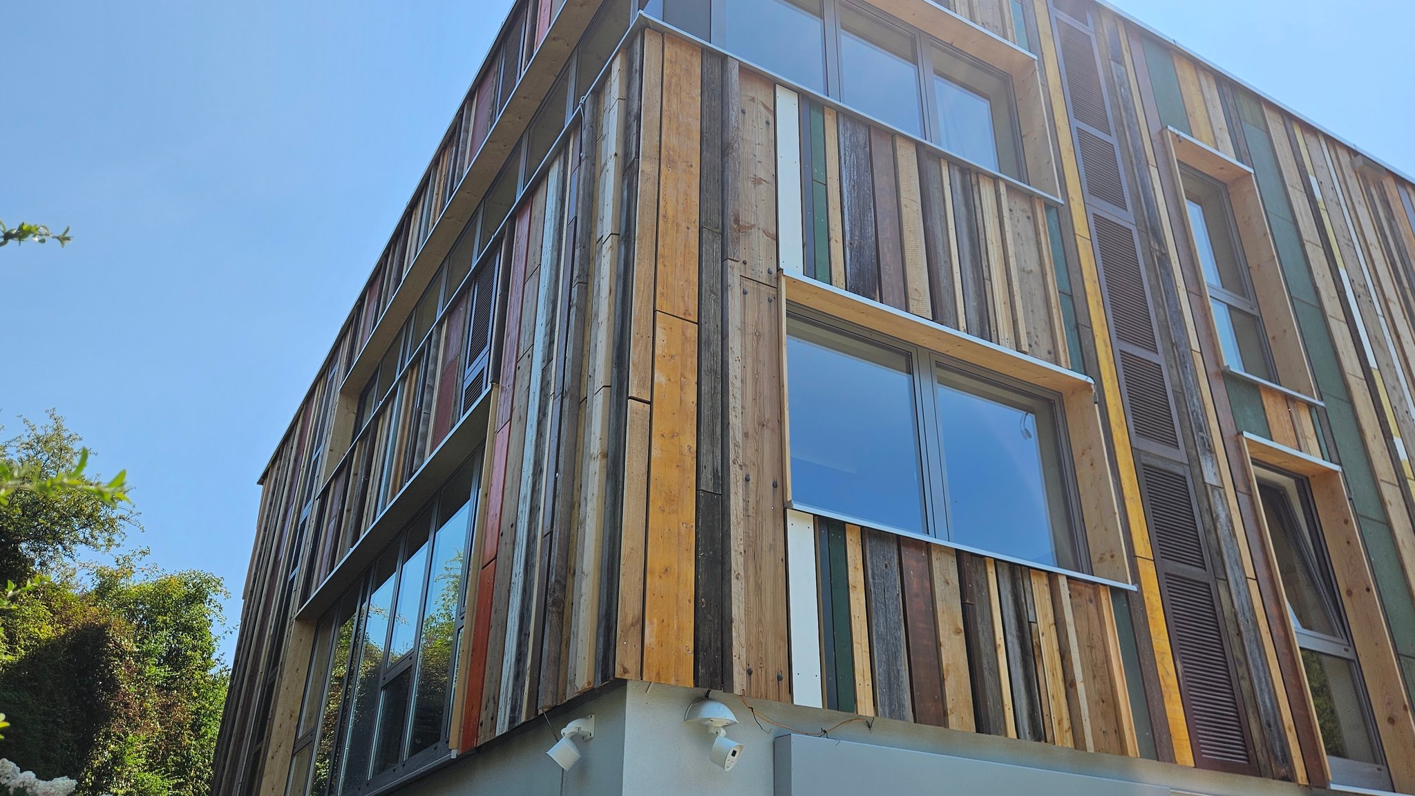 Ein Gebäude mit einer Holzfassade. Die Bretter sind senkrecht angeordnet. Sie sind nicht nur holzfarben, sondern zum Teil auch grün, gelb, braun, weiß und haben eine unterschiedliche Struktur