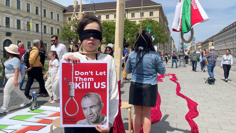Eine Demonstrantin hält ein Schild hoch: Don't Let Them Kill US". Sie steht an einem Galgen, hinter ihr markieren zwei rote Stoffbahnen eine lange Blutspur.