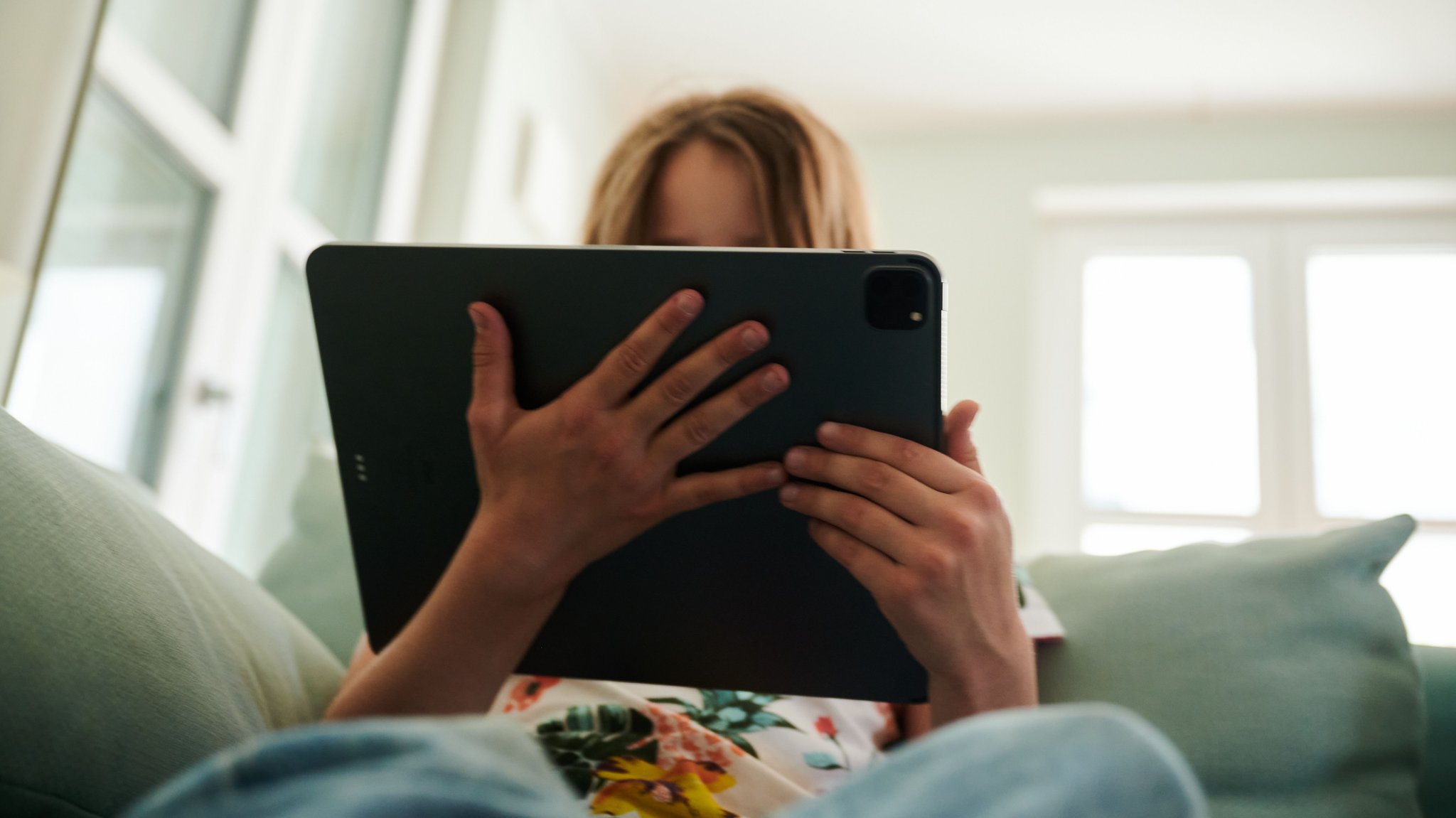 (Symbolbild) Ein Mädchen sitzt auf der Couch und guckt in das Tablet ihrer Eltern. 