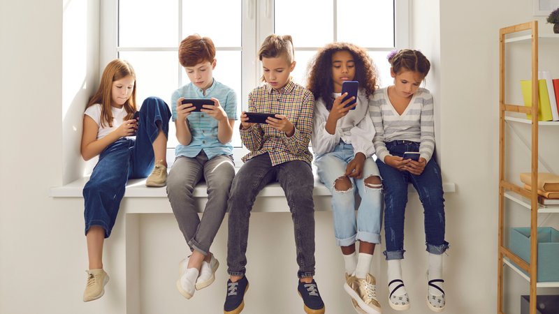 Fünf Kinder diverser Nationalitäten an ihren Handys auf einer Fensterbank. | Bild: stock.adobe.com/Studio Romantic Fünf Kinder diverser Nationalitäten an ihren Handys auf einer Fensterbank.