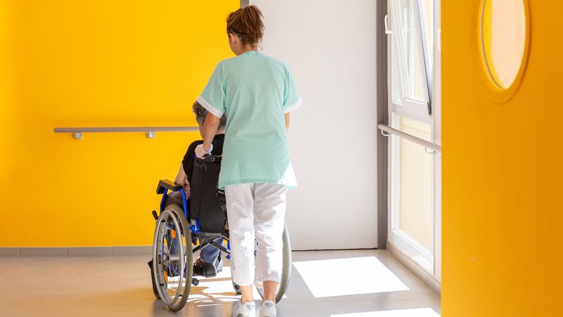 Eine Altenpflegerin schiebt eine im Rollstuhl sitzende Person.