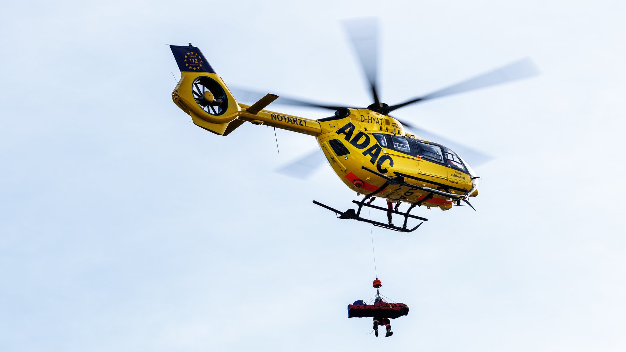 (Symbolbild) Ein Luftretter vom ADAC hängt bei einer Vorführung mit einem Bergesack an der Winde von einem Rettungshubschrauber vom Typ Airbus H145. 