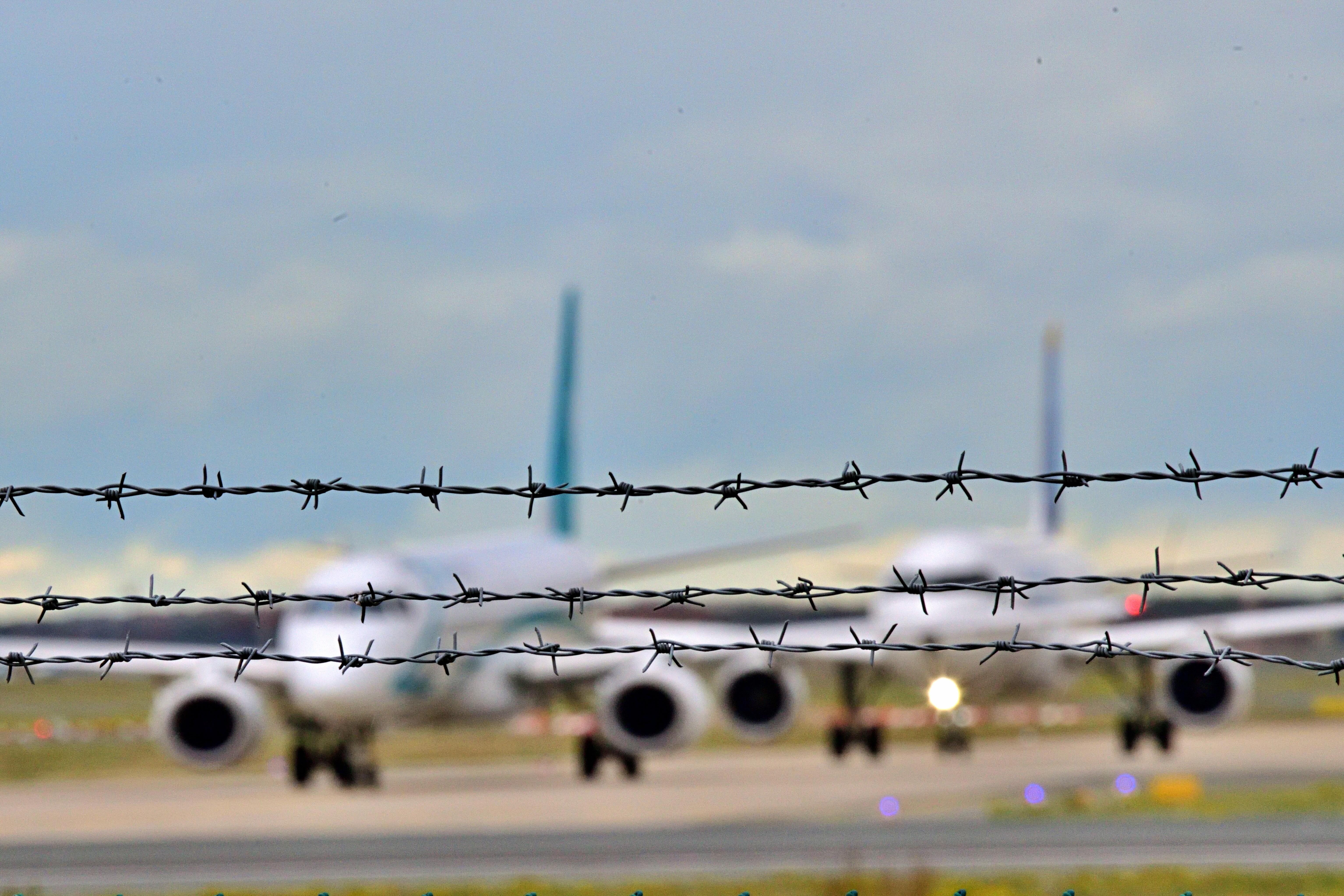 Drei Reihen Stacheldraht, dahinter in Unschärfe zwei Flugzeuge auf der Landebahn. (Symbolbild) Der Bügerkrieg in Syrien ist beendet. Soll in das Land wieder abgeschoben werden? Über diese Frage ist eine Debatte entbrannt. 