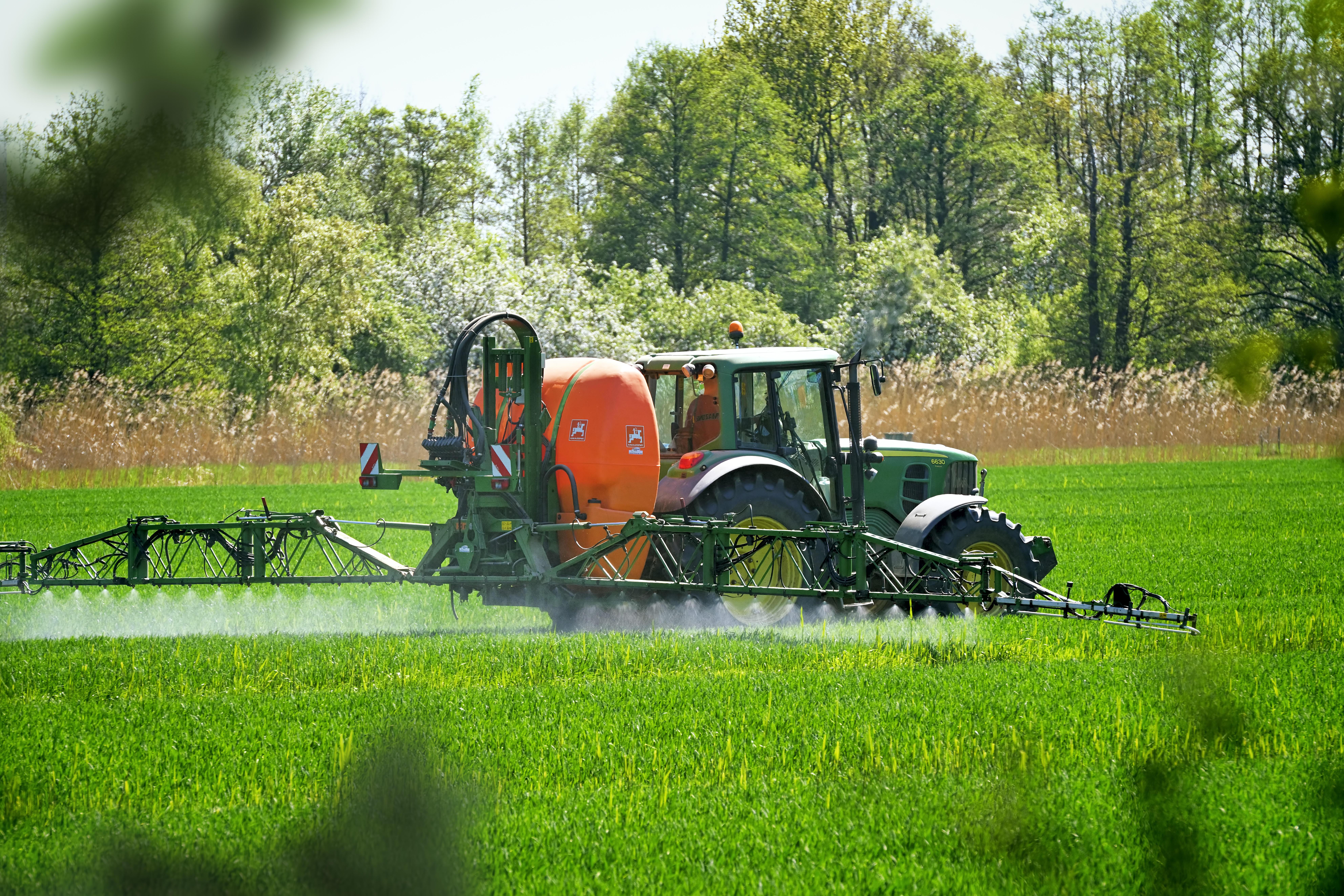 (Symbolbild) Grüner Traktor mit orange-farbener Feldspritze fährt über ein Getreidefeld