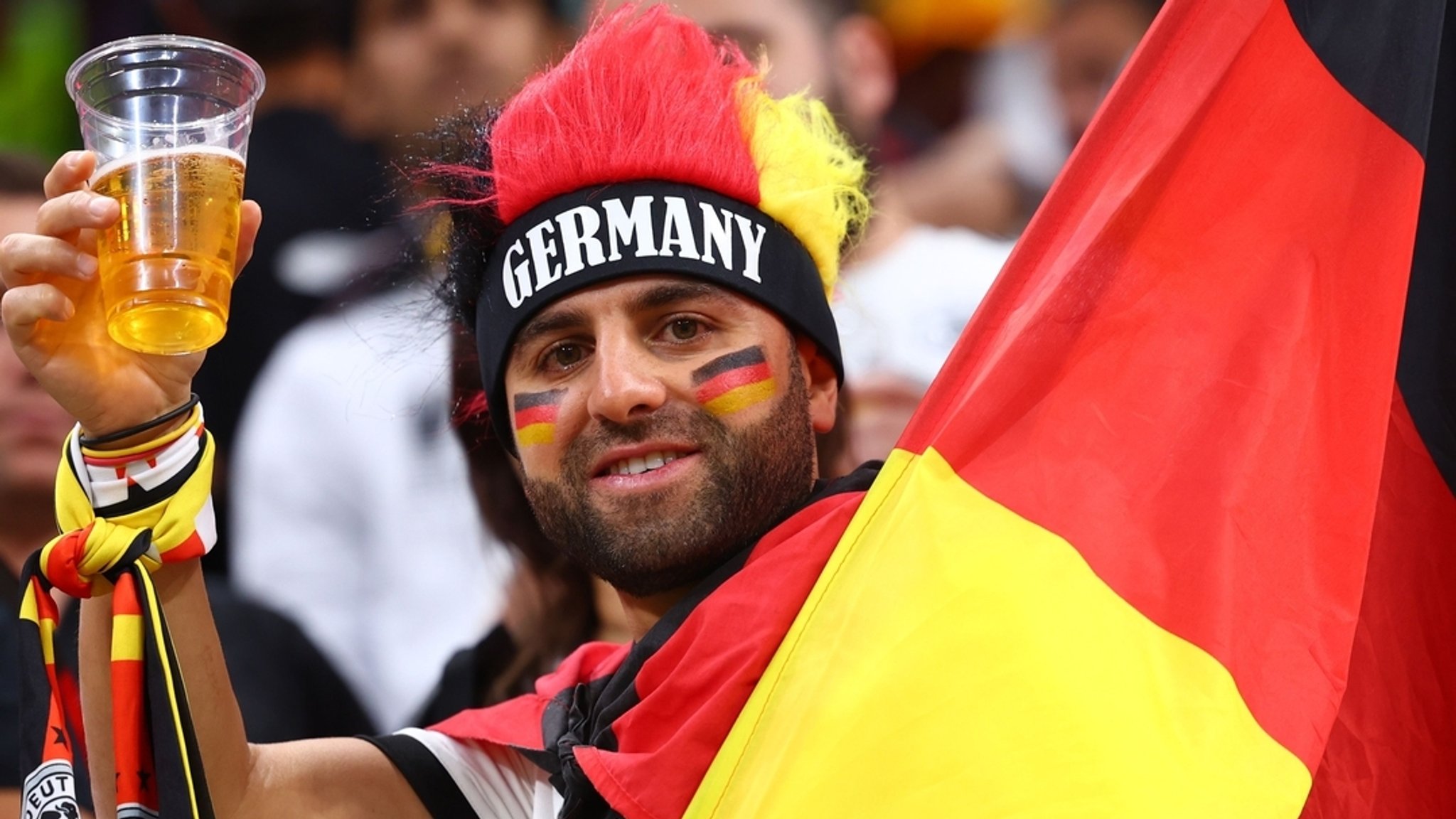 Ein Fan der deutschen Mannschaft lacht vor einem Spiel bei der WM 2022 in Katar und hält einen halb leeren Bierbecher in der Hand.