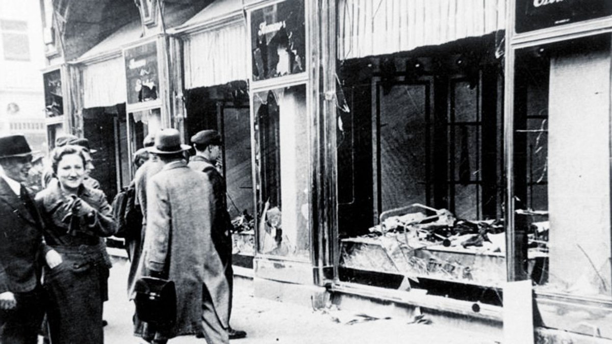 1938: Passanten gehen an zerstörten jüdischen Geschäften vorbei. 