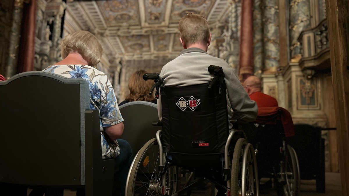 Im Opernhaus Bayreuth sitzen Menschen im Parkett. Neben der Stuhlreihe sitzt ein Mann im Rollstuhl. 