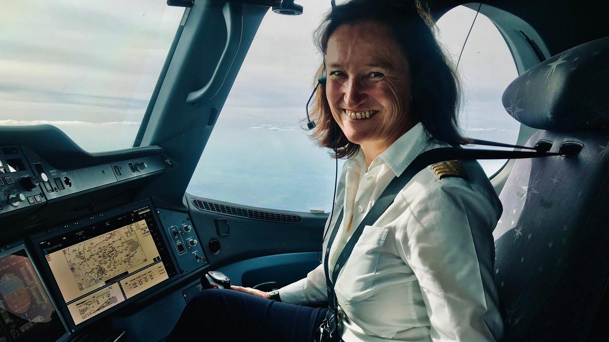 Eine Pilotin sitzt angeschnallt im Cockpit und lächelt. Vor ihr befindet sich ein Bildschirm und aus dem Fenster heraus sieht man den Himmel.