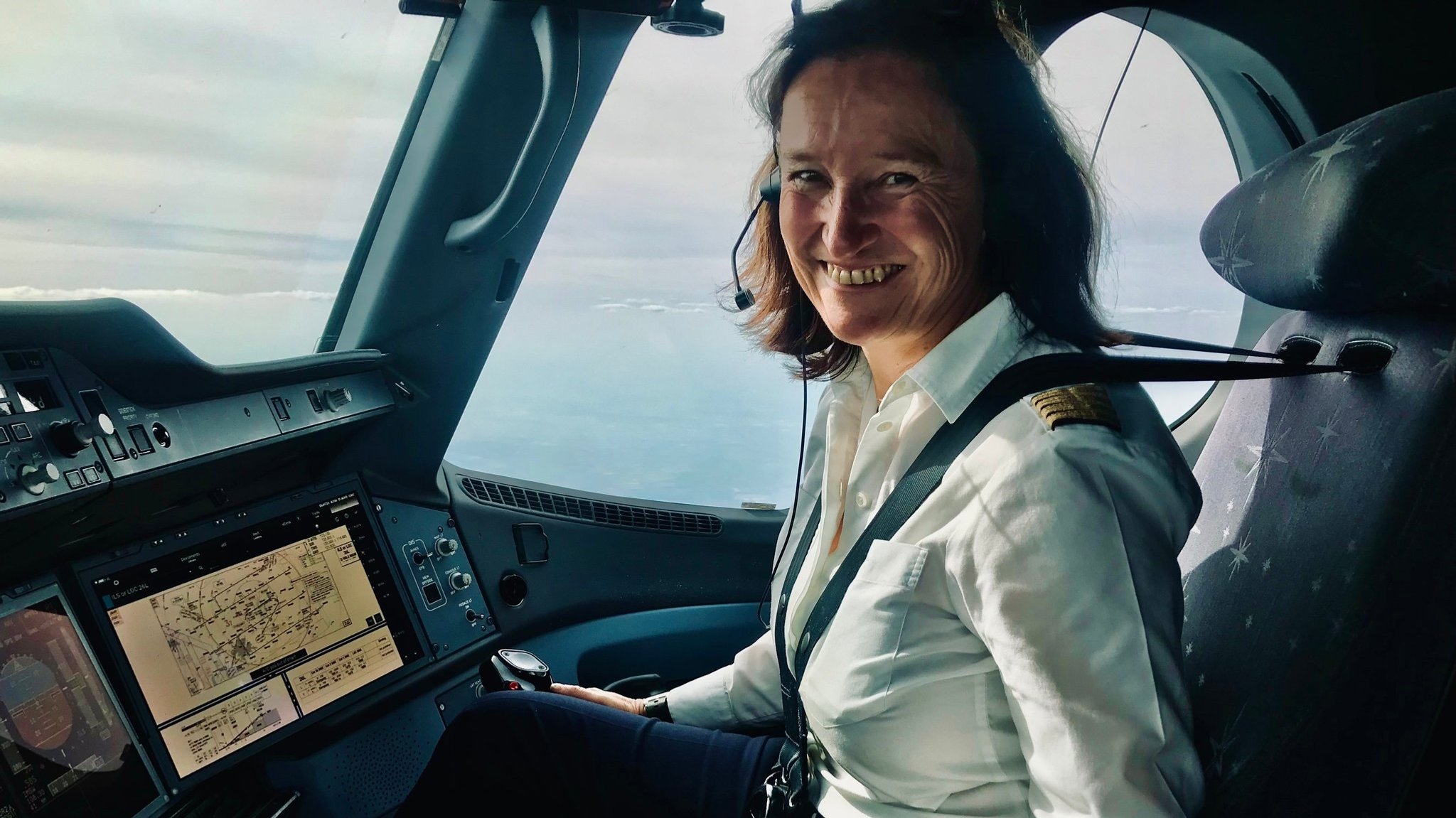 Eine Pilotin sitzt angeschnallt im Cockpit und lächelt. Vor ihr befindet sich ein Bildschirm und aus dem Fenster heraus sieht man den Himmel.