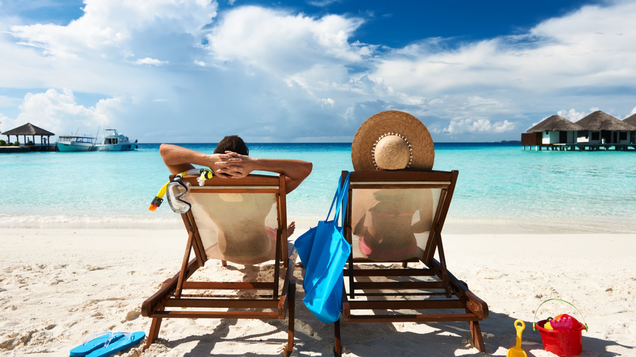 Ein Pärchen sitzt in Liegestühlen am Strand und blickt auf das hellblaue Meer vor sich.