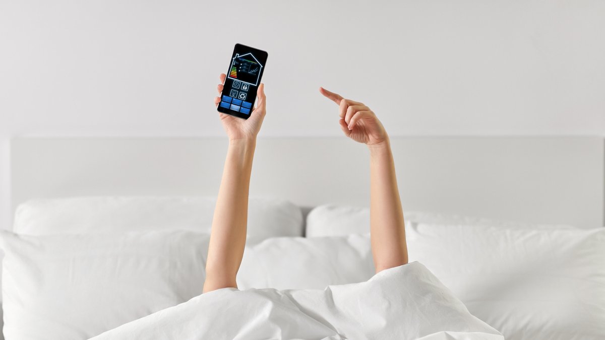 Frau liegt im Bett mit Smartphone, auf dem Smart Home-Icons zu sehen sind (Symbolbild)