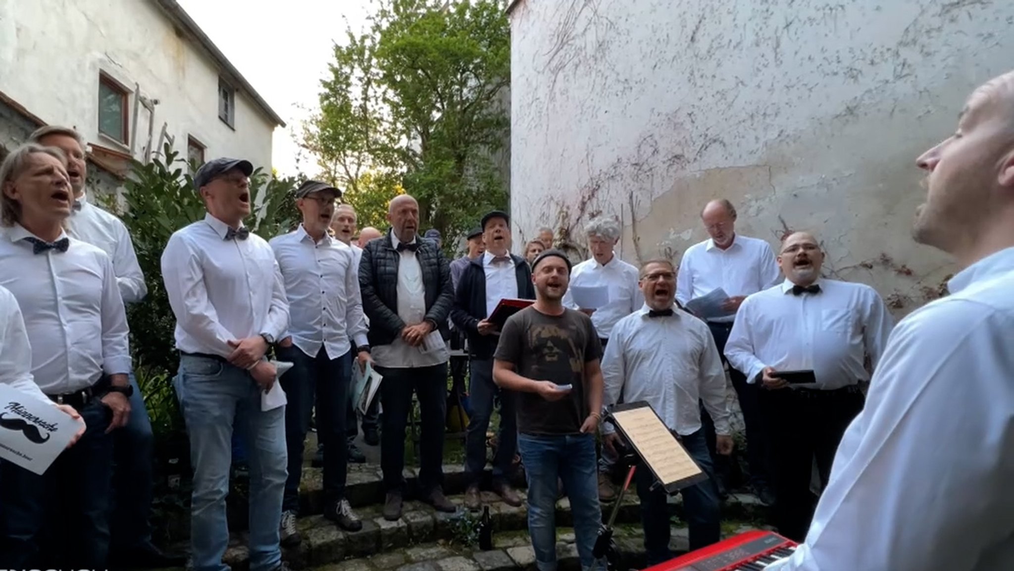 Mehrere Männern mit weißen Hemden und Fliege singen, einige halten Liederblätter in der Hand. 