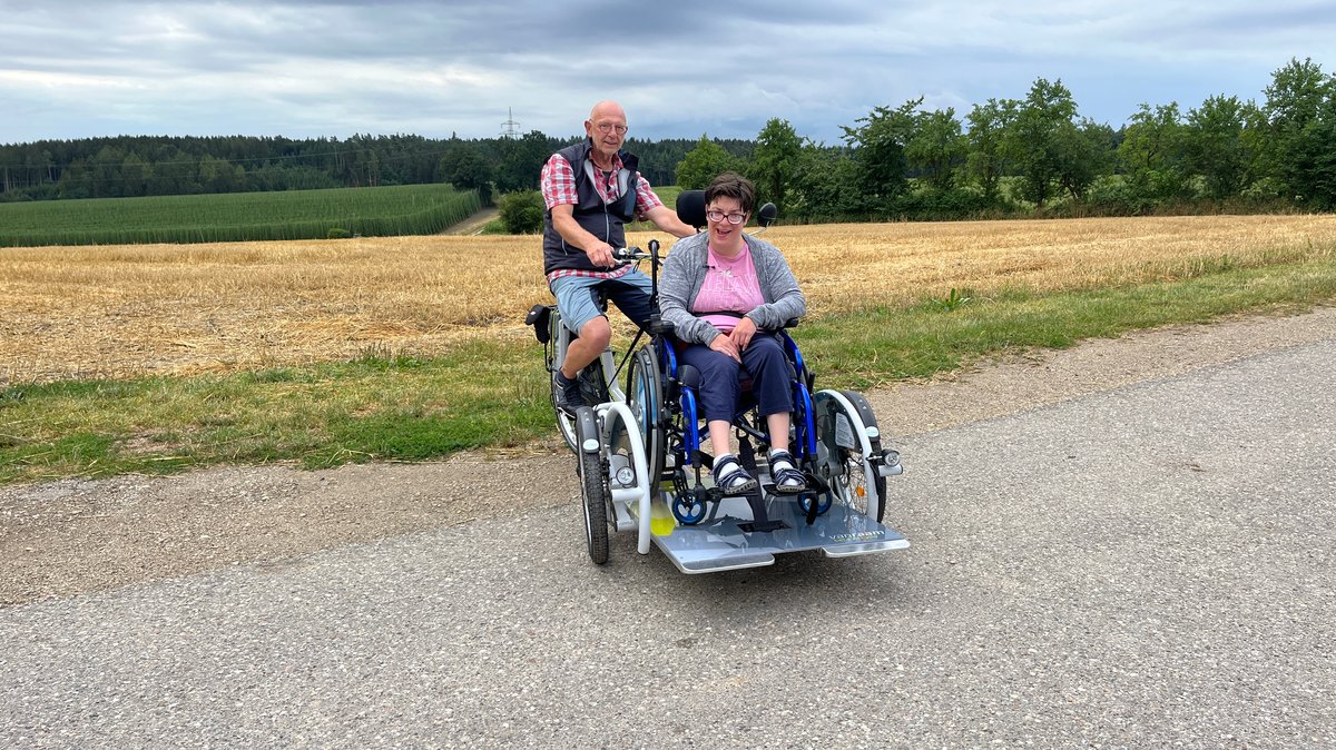 Eine Frau im Rollstuhl und ein Mann im Rentenalter auf dem Fahrradsitz lächeln in die Kamera. Der Rollstuhl ist auf einer Plattform zwischen den beiden Vorderrädern der Rolli-Rikscha befestigt.