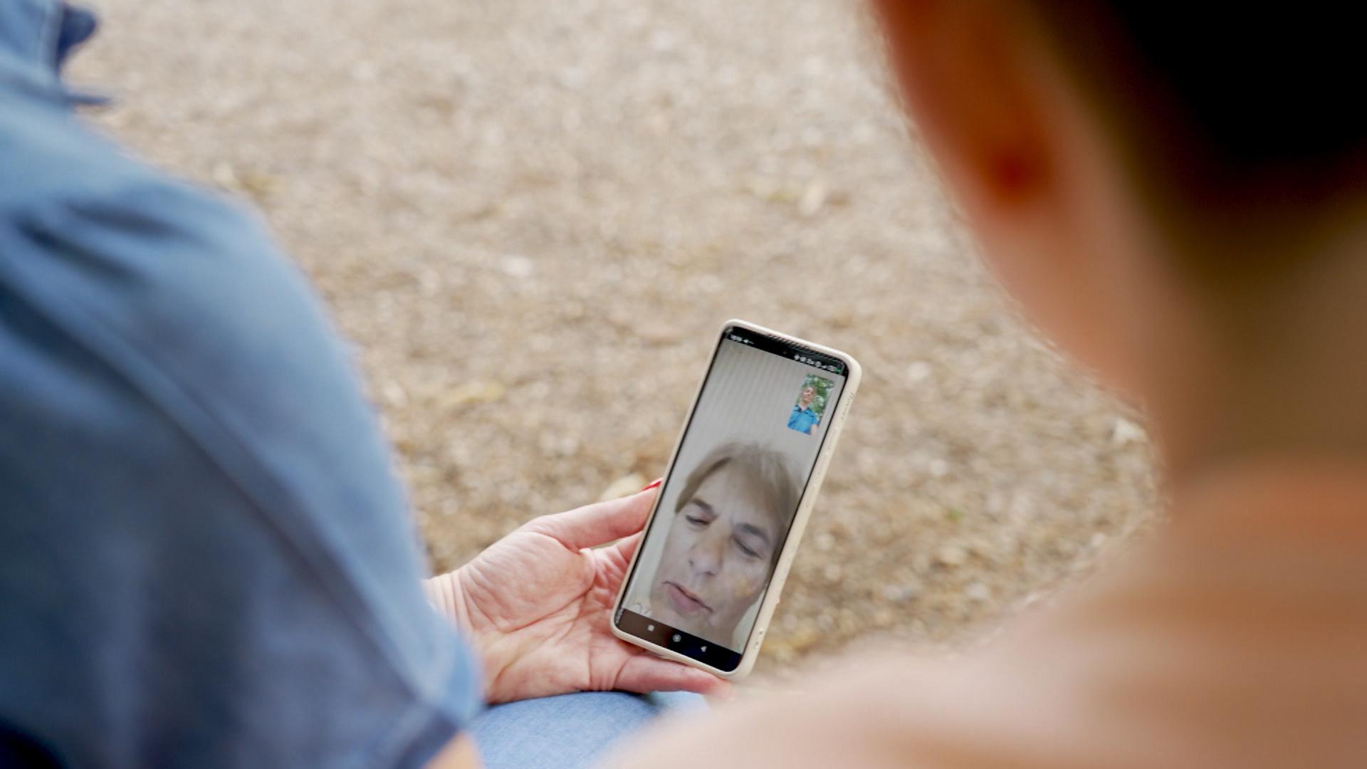 Eine Frau hält ein Handy auf dem ein Videoanruf mit ihrer Mutter zu sehen ist.
