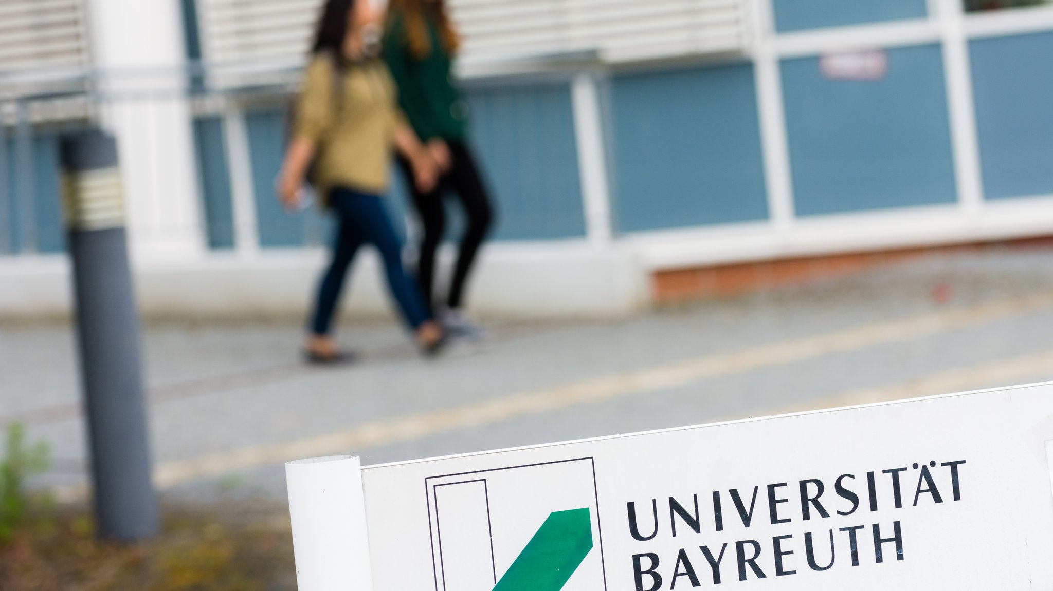 Auf einem Schild steht "Universität Bayreuth". Im Hintergrund laufen zwei Studentinnen. 