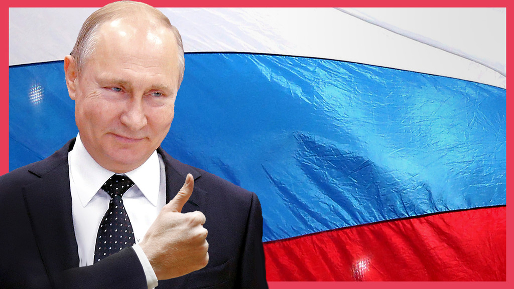 Ein Mann in Anzug und Krawatte, es ist der russische Präsident Wladimir Putin, vor ihm kommt eine Hand, die die Daumen nach oben Geste macht ins Bild, hinter ihm die russische Flagge: weiß, blau, rot.