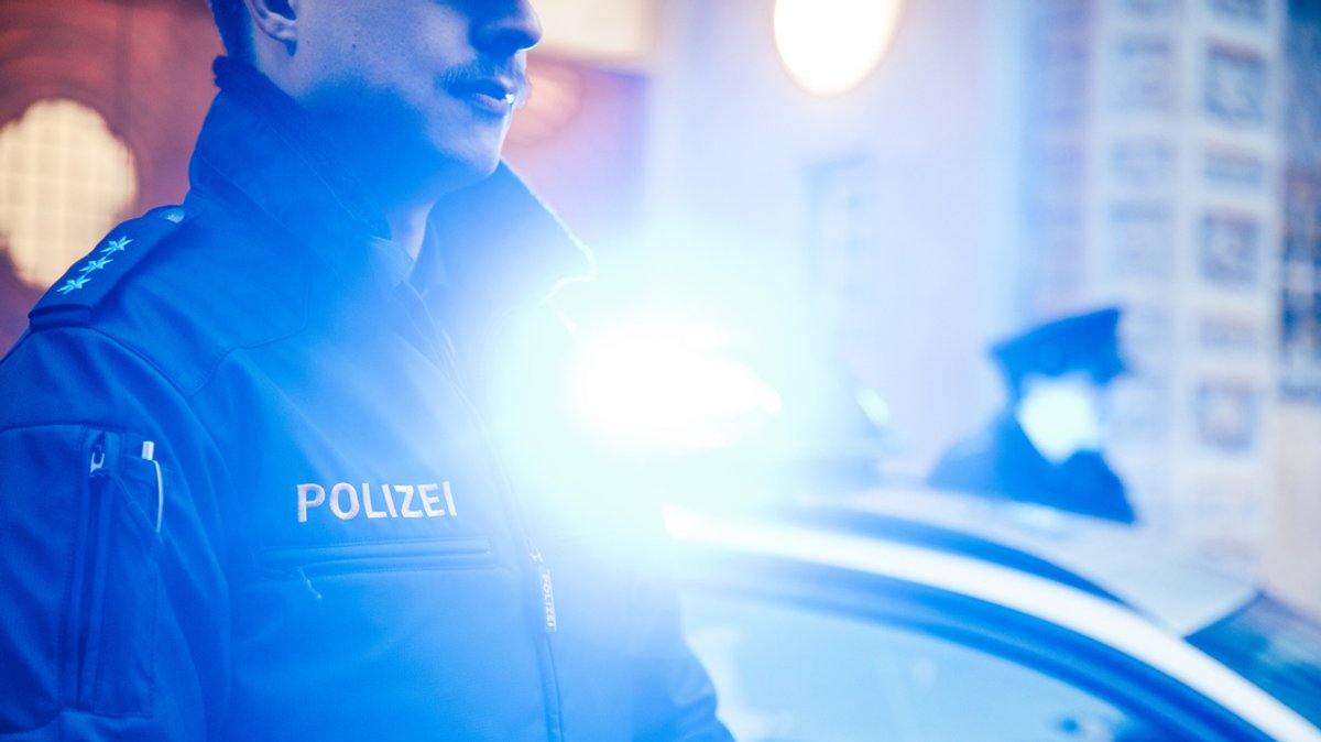 Eine Polizistin und ein Polizist stehen vor dem Eingang des Polizeipräsidiums München vor einem Polizeiauto mit Blaulicht.