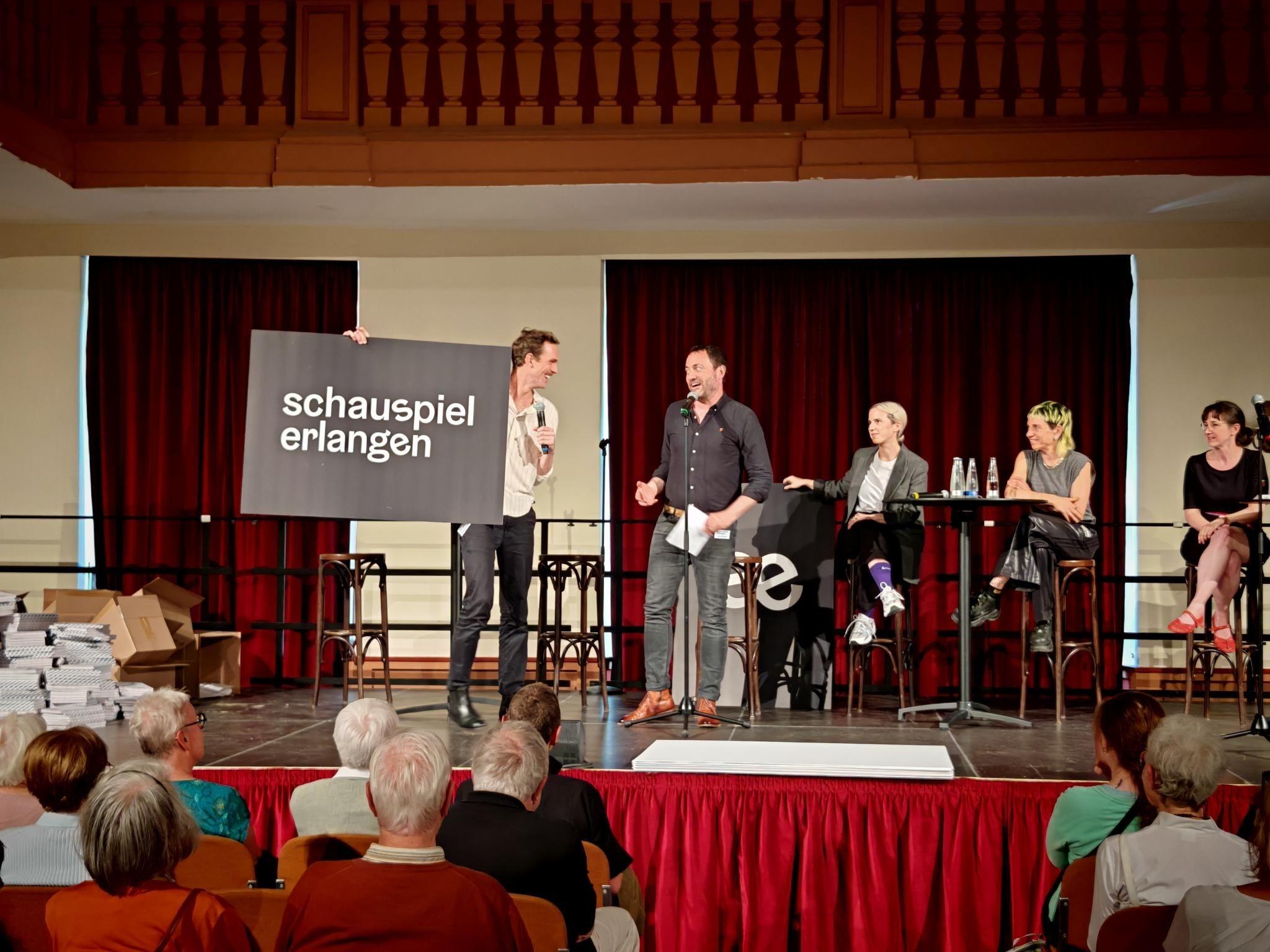 Ein Schild mit der Aufschrift "Schauspiel Erlangen" vor einem roten Theatervorhang.