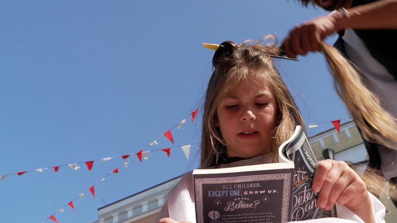 Ein Mädchen liest in einem Buch. Währenddessen werden ihr die Haare geschnitten. | Bild: BR/Melina Eisemann Ein Mädchen liest in einem Buch. Währenddessen werden ihr die Haare geschnitten.