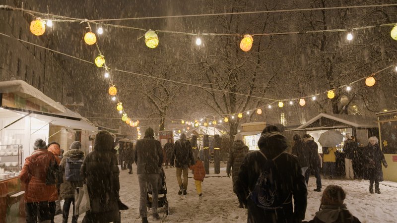 Menschen gehen im Schneegestöber zwischen den Buden auf dem Schwabinger Weihnachtsmarkt in München umher | Bild: BR/megaherz gmbh Menschen gehen im Schneegestöber zwischen den Buden auf dem Schwabinger Weihnachtsmarkt in München umher