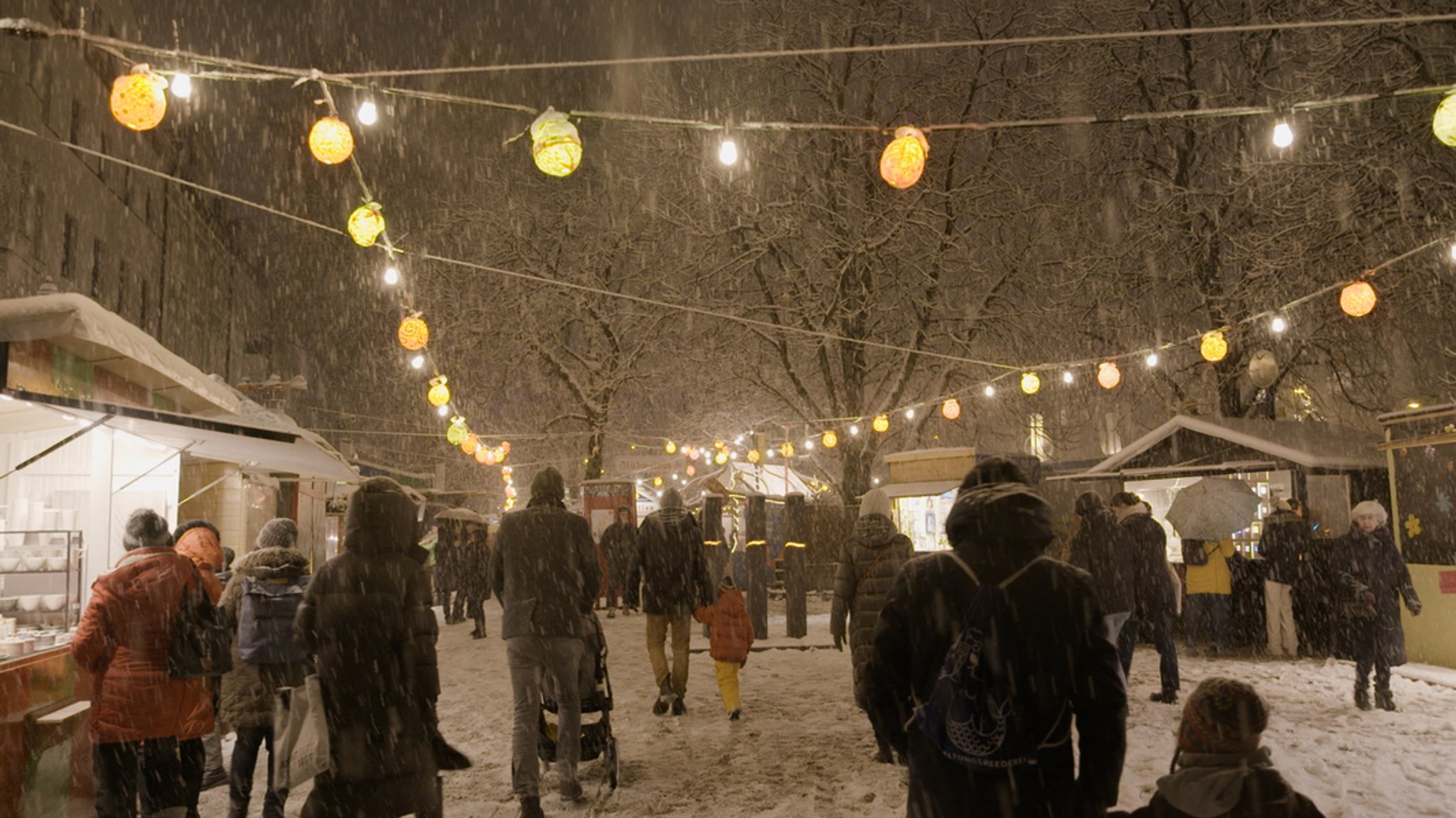 Menschen gehen im Schneegestöber zwischen den Buden auf dem Schwabinger Weihnachtsmarkt in München umher