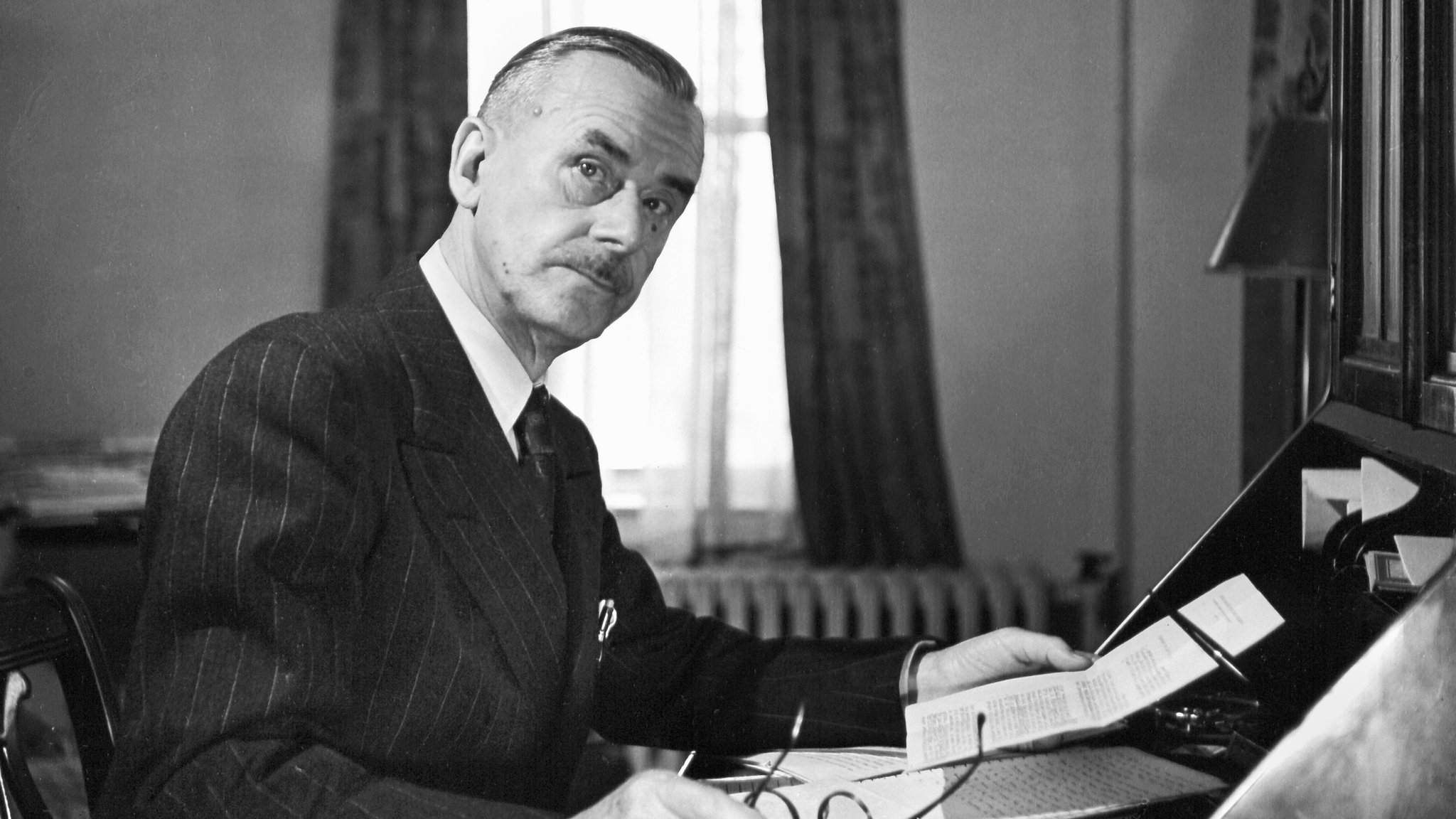 Der Schriftsteller Thomas Mann im Exil in den USA im Jahr 1943, an seinem Schreibtisch sitzend, in einer Hand seine Brille, in der anderen ein Schriftstück