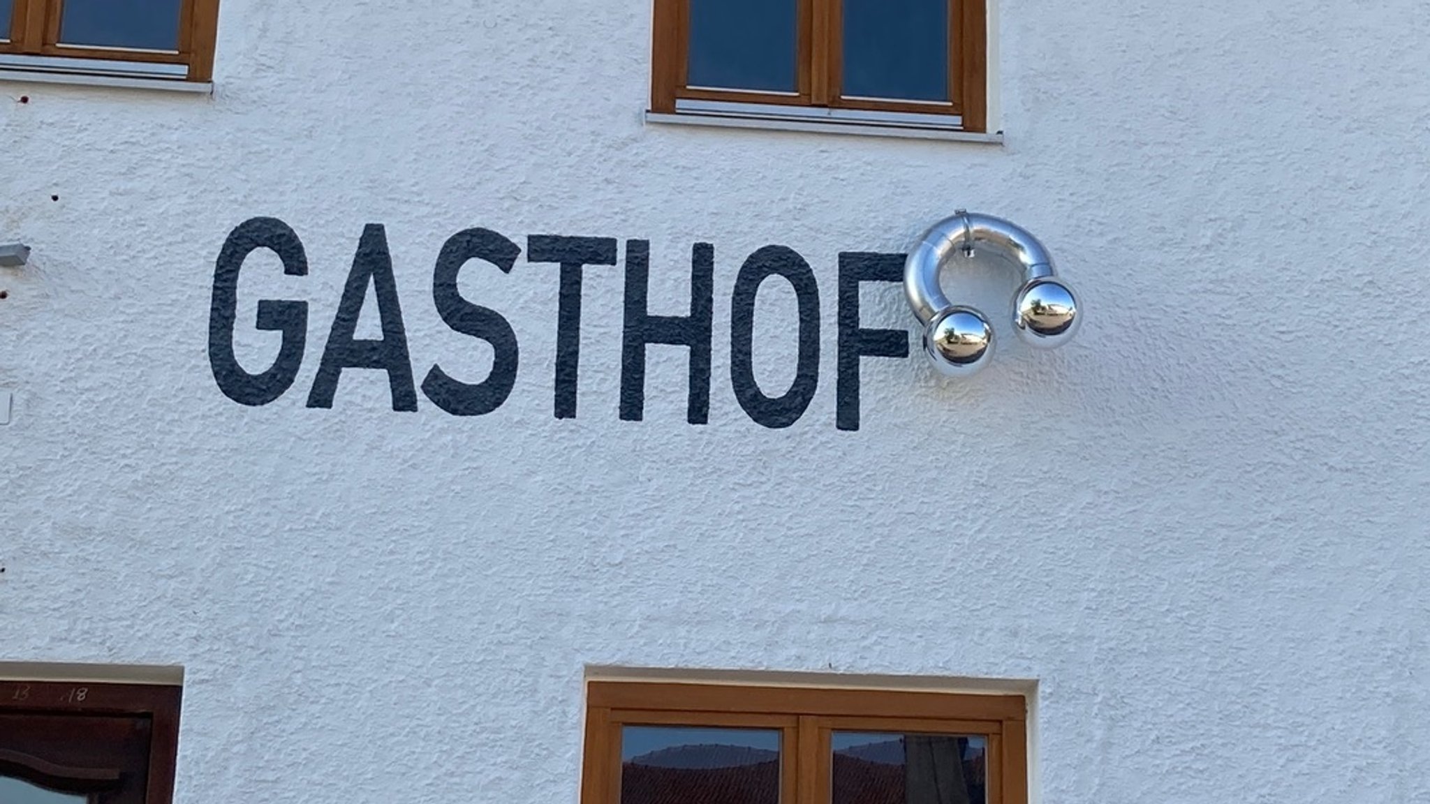 Eine Hausfassade mit der Aufschrift "Gasthof", an dessen Ende ein überdimensioniertes Piercing baumelt.