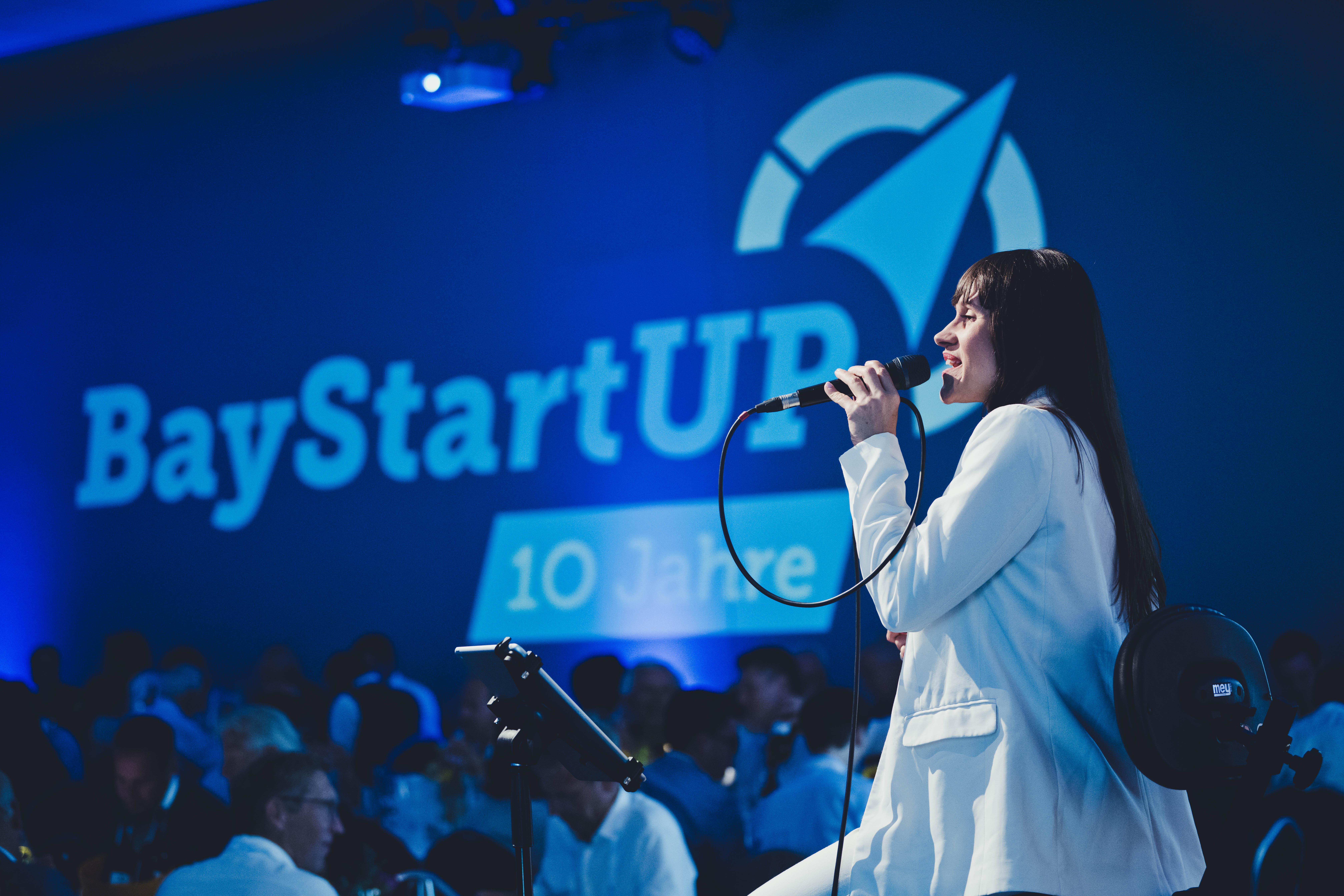 Eine junge Frau steht auf einer Bühne, im Hintergrund der Schriftzug BayStartUp