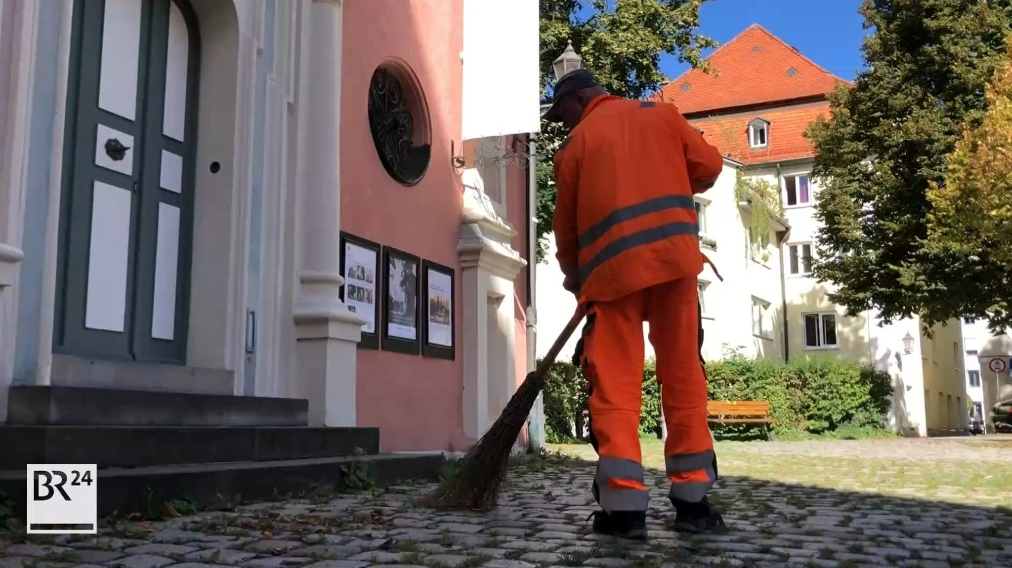 Ein Mensch in oranger Straßenreinigungskluft von hinten. Er fegt die Straße.