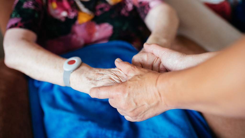 Eine jüngere Frau hält die Hände einer älteren Frau, die einen Notrufknopf am Handgelenk hat.