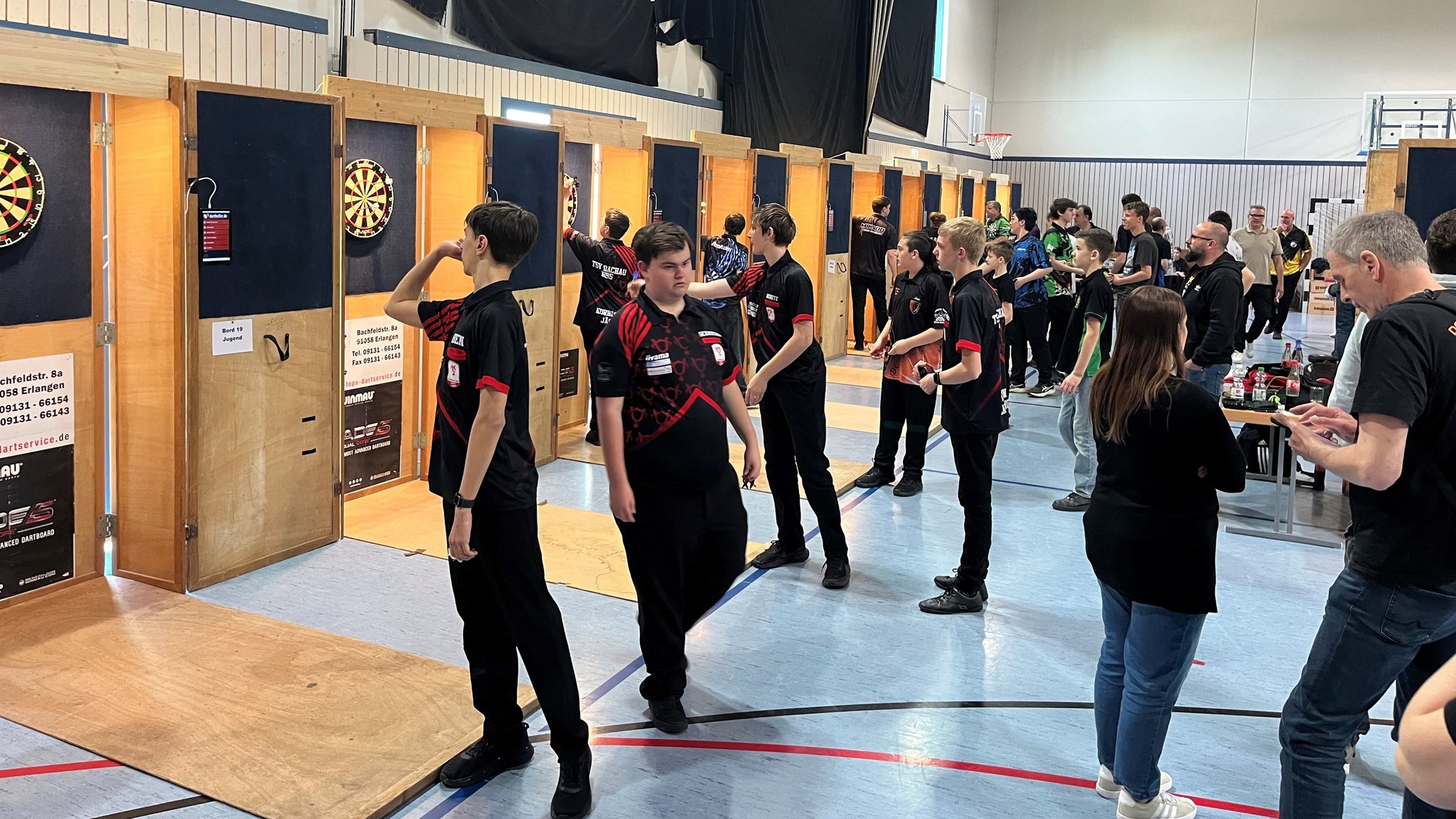Mehrere Dartboards, im Vordergrund vor allem jugendliche Werfer beim Einwerfen. 