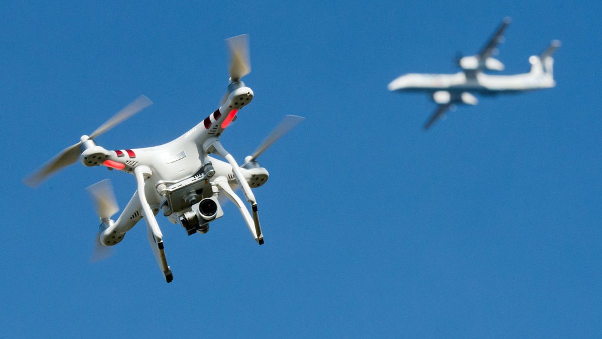 Eine Drohne fliegt umher, während im Hintergrund am Himmel ein Flugzeug unterwegs ist (Archiv)