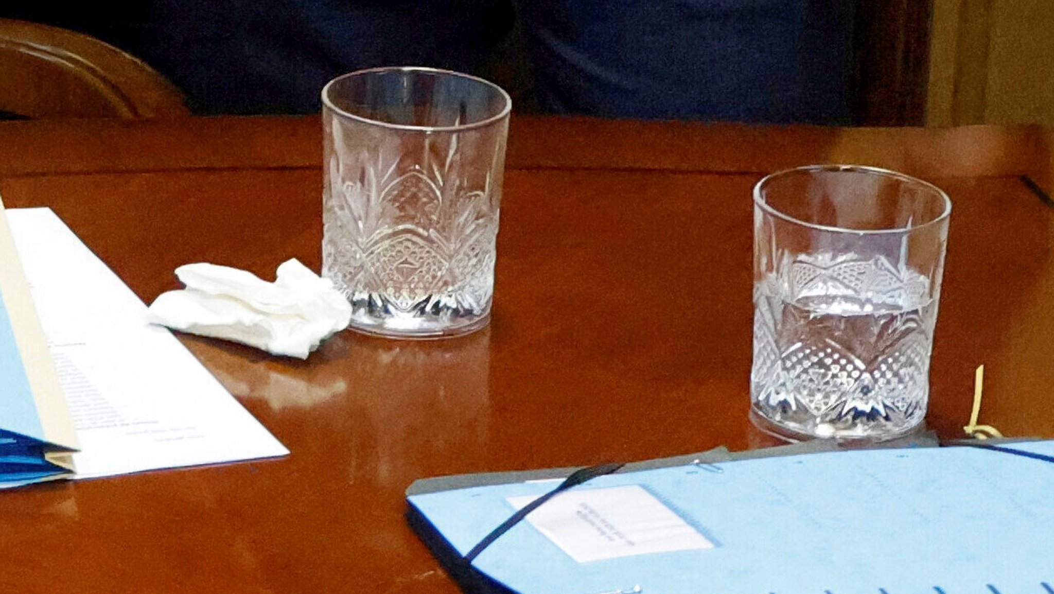 Zwei Wassergläser, Aktenordner, ein weißes Taschentuch und ein Stäbchen stehen oder liegen auf einer Tischplatt.