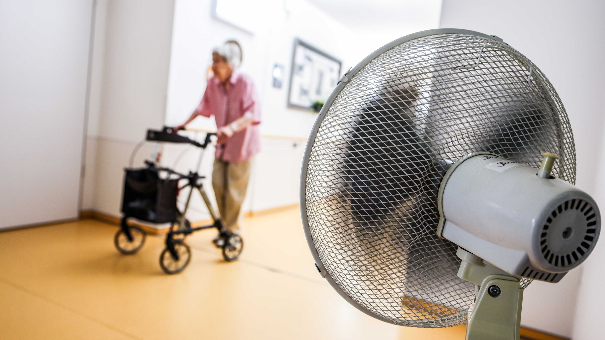 Symbolbild: Eine Frau geht mit einem Rollator in einem Seniorenheim über einen Flur, der aufgrund hoher Außentemperaturen mit einem Ventilator belüftet wird.