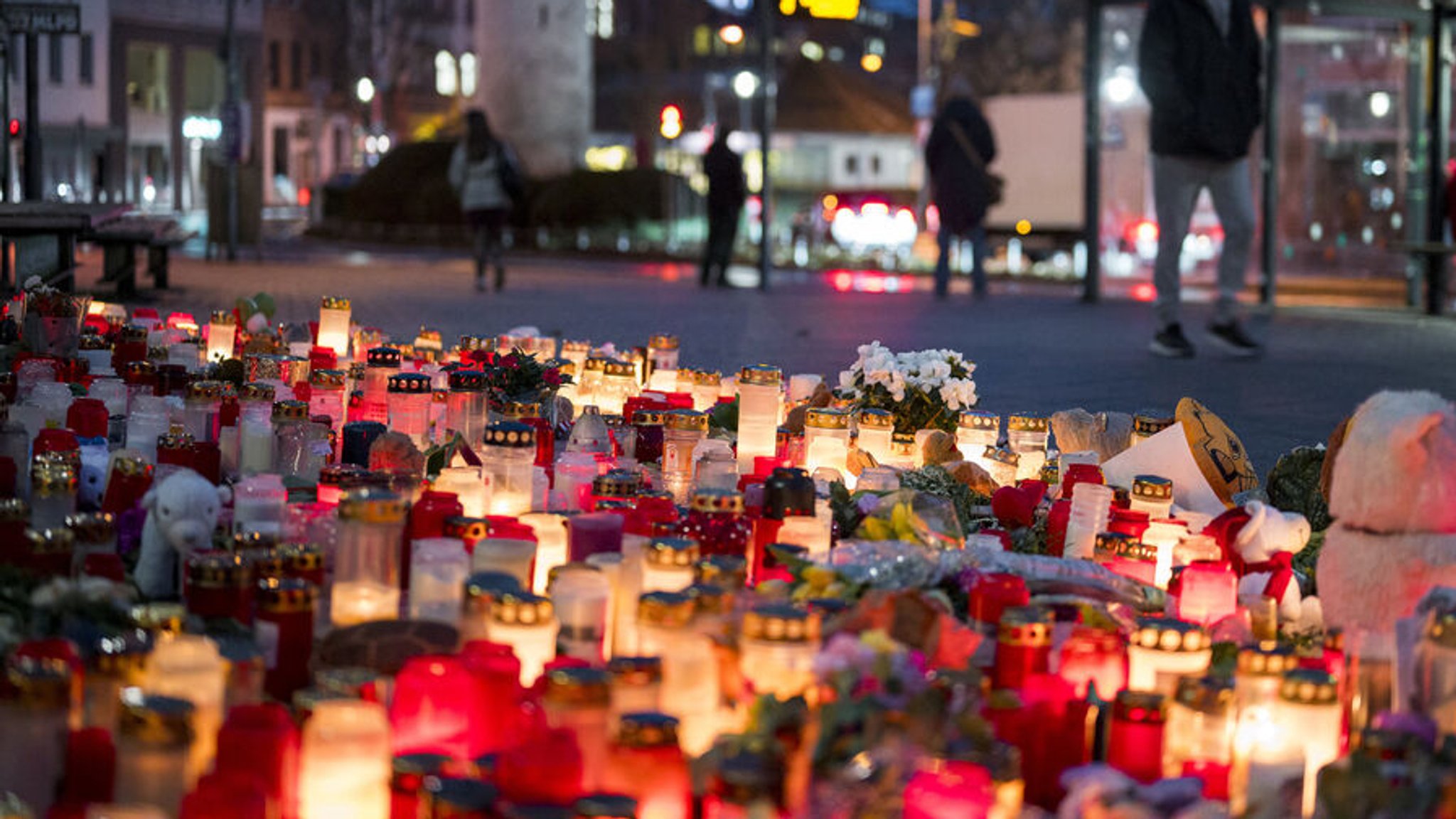 27.01.2025, Bayern, Aschaffenburg: Zahlreiche Kerzen und Blumen stehen am Eingang des Parks Schöntal. Im Hintergrund warten Personen an einer Bushaltestelle. Foto: Daniel Vogl/dpa +++ dpa-Bildfunk +++