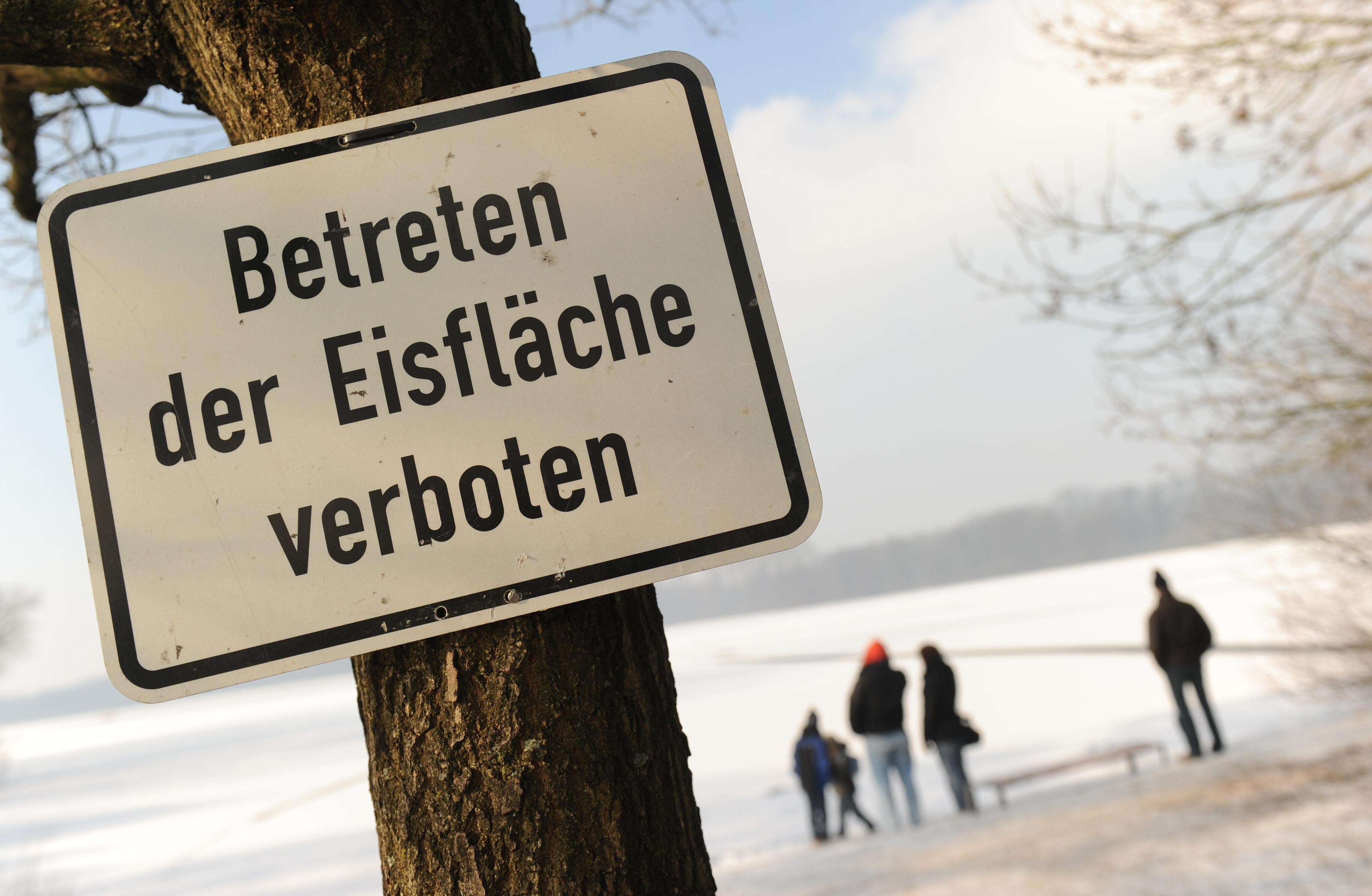 Archivbild: Ein Schild mit der Aufschrift: "Betreten der Eisfläche verboten", dahinter ein zugefrorener See mit Menschen, die am Ufer stehen.