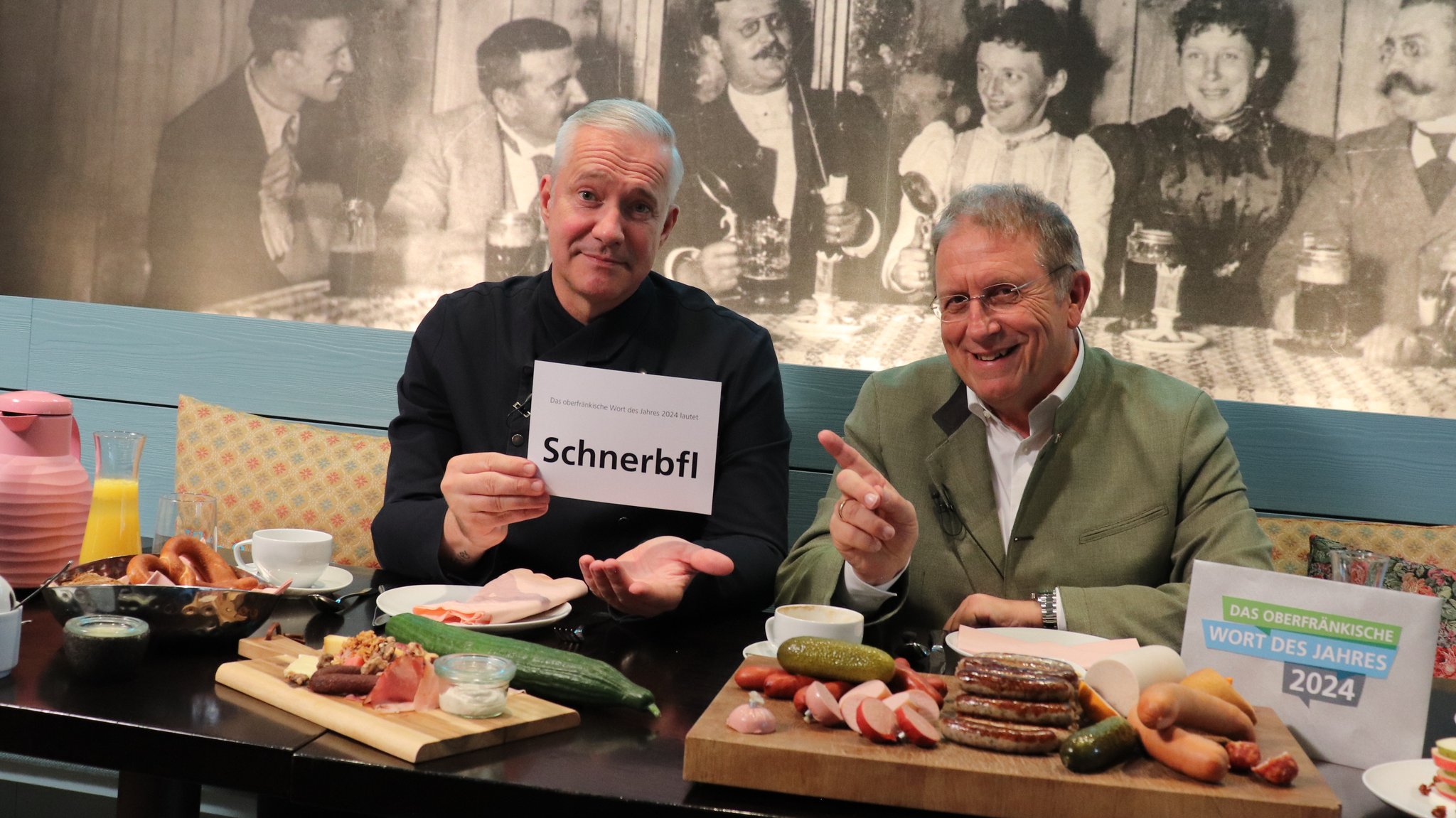 Alexander Herrmann und Henry Schramm sitzen vor Platten mit Wurst. Alexander hält ein Schild mit dem Wort Schnerbfl hoch.