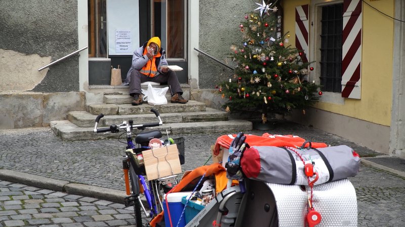 Mann sitzt auf Steinstufen vor Haus, davor bepacktes Fahrrad