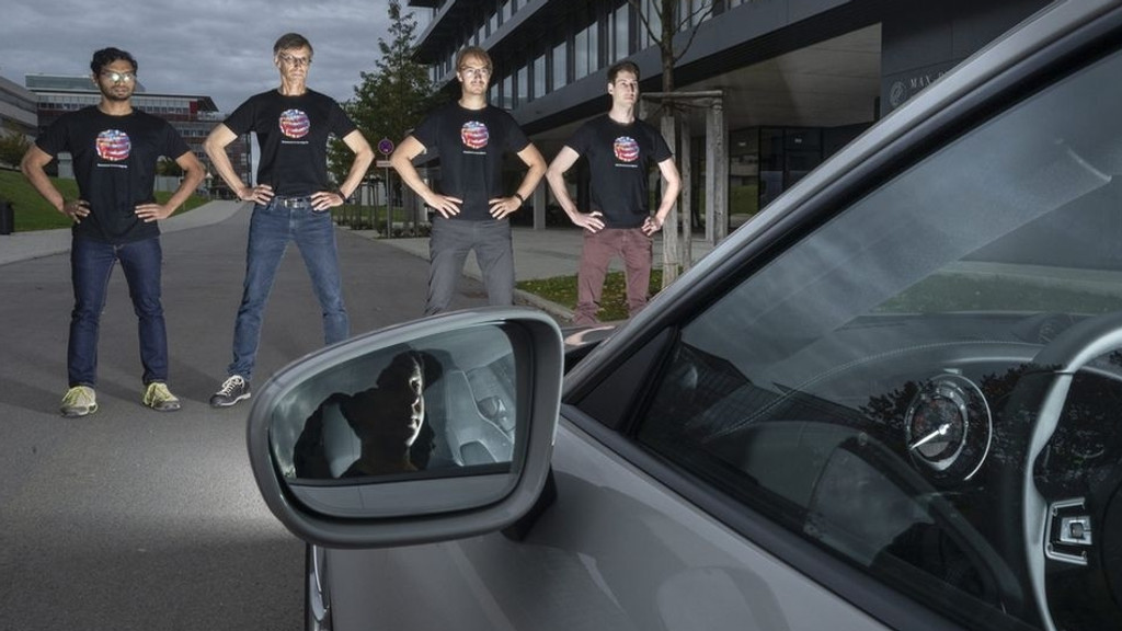 Die Wissenschaftler Anurag Ranjan (von links nach rechts), Michael J. Black, Andreas Geiger und Joel Janai stehen mit Farbflecken bedruckten T-Shirts vor einem Auto.