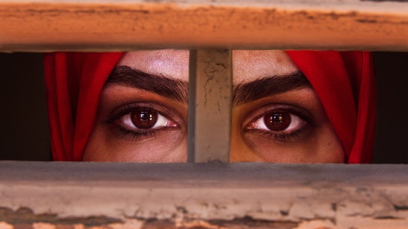 Liza Anvary, eine afghanische Geflüchtete steht hinter einem Holzfenster, als befände sie sich im Gefängnis - um, die Situation der Frauen in ihrem Land darzustellen.